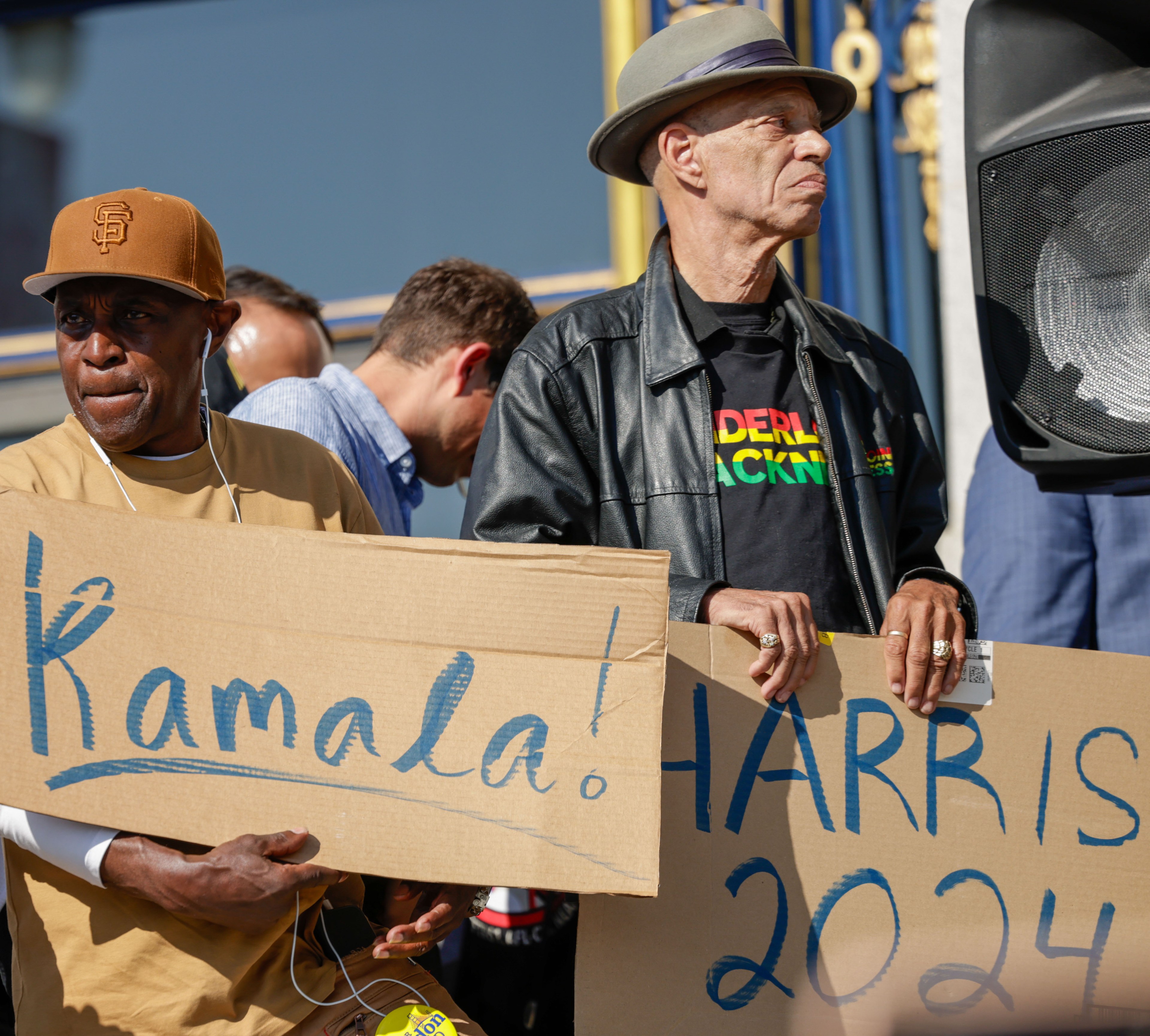 Two men hold signs reading &quot;Kamala!&quot; and &quot;Harris 2024.&quot; One wears a tan hat and shirt, the other a fedora and leather jacket. A large speaker and other people are in the background.