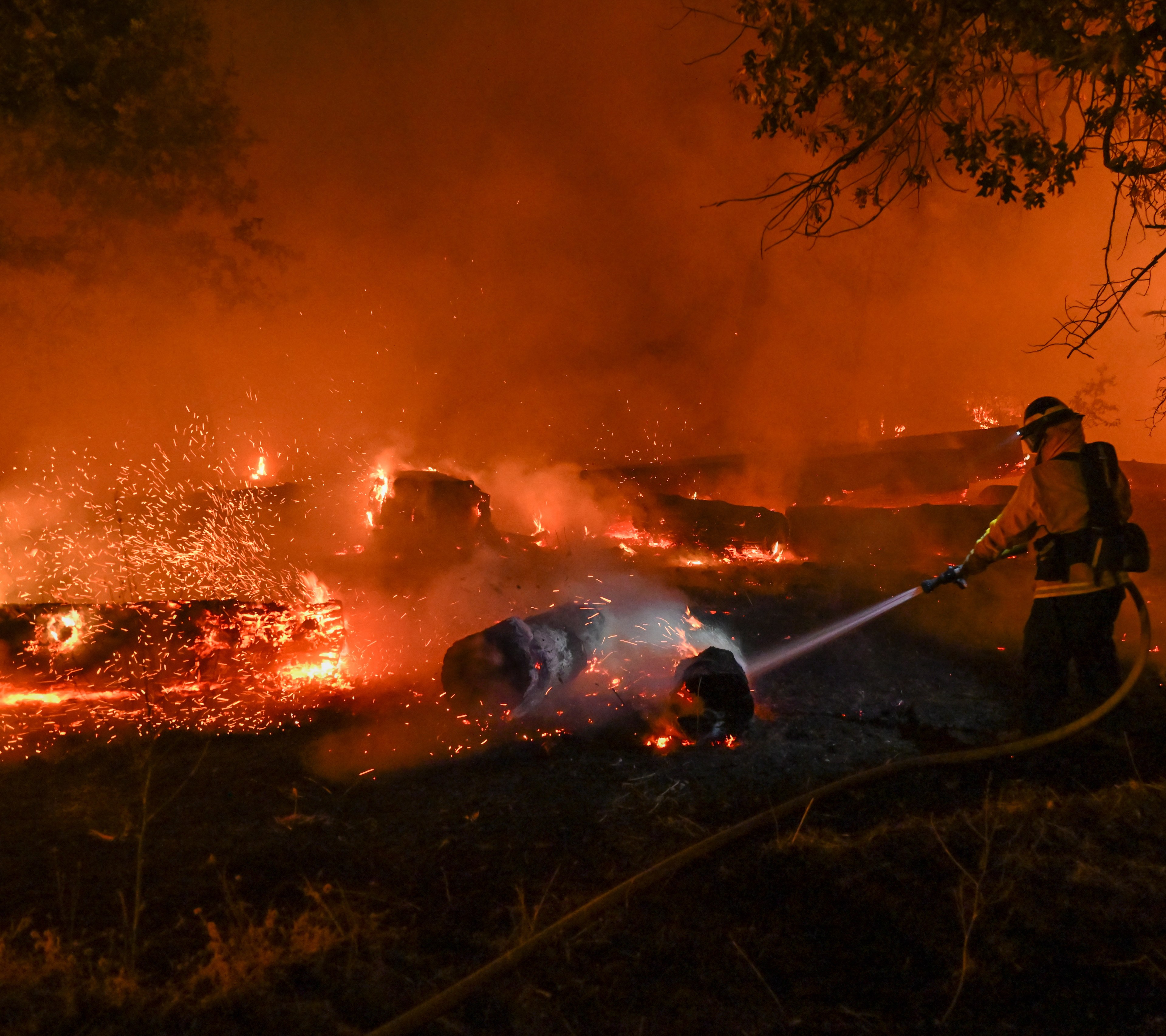 A firefighter uses a hose to combat a large, intense wildfire. The landscape is engulfed in flames and heavy smoke, with glowing embers scattered around.