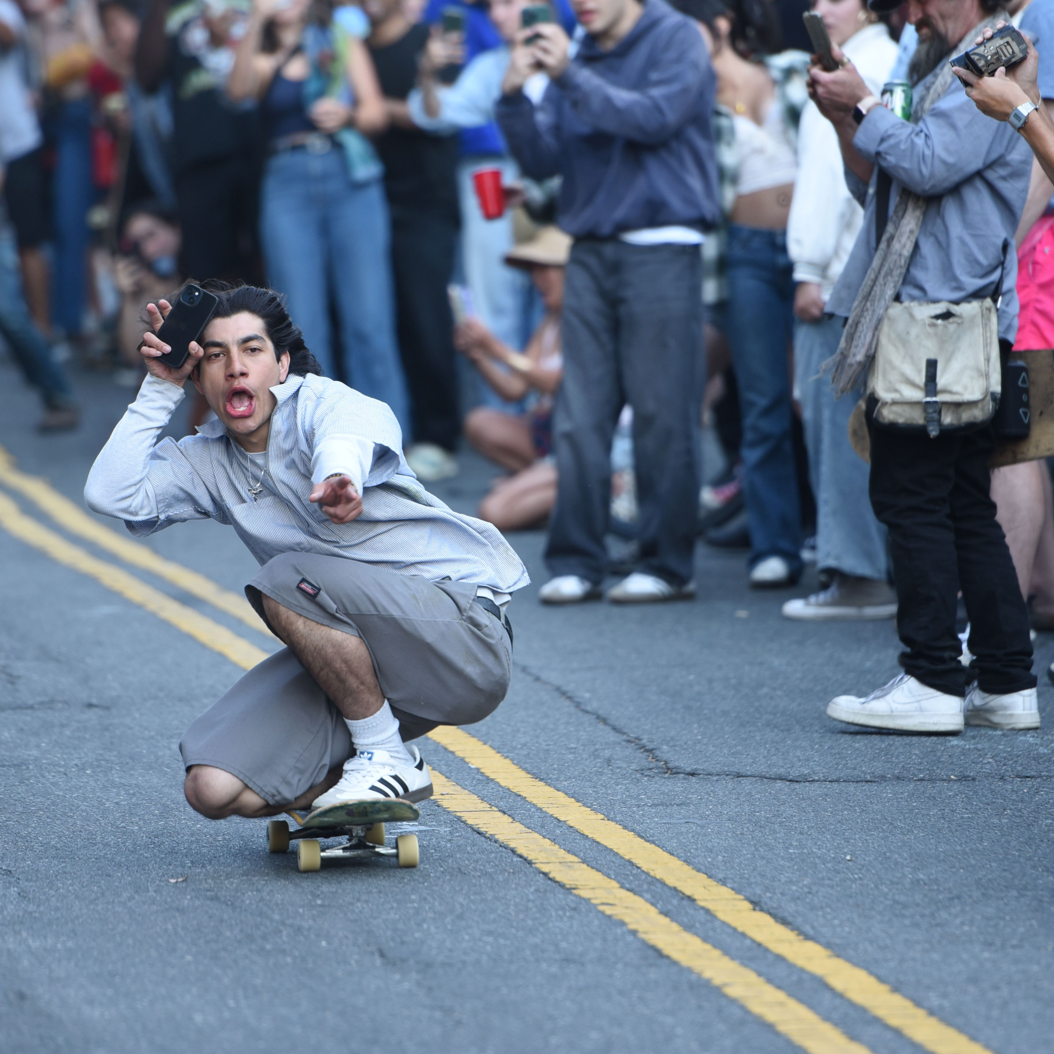 A skater in a gray shirt and shorts rides a skateboard down a street, holding a phone. He's surrounded by a crowd capturing the moment with their phones.