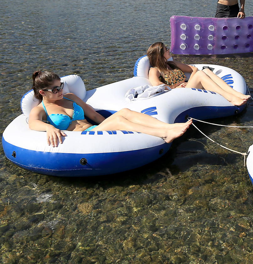 Two women in bikinis relax on a large blue and white inflatable float in a shallow body of water. A purple raft and another person are visible in the background.