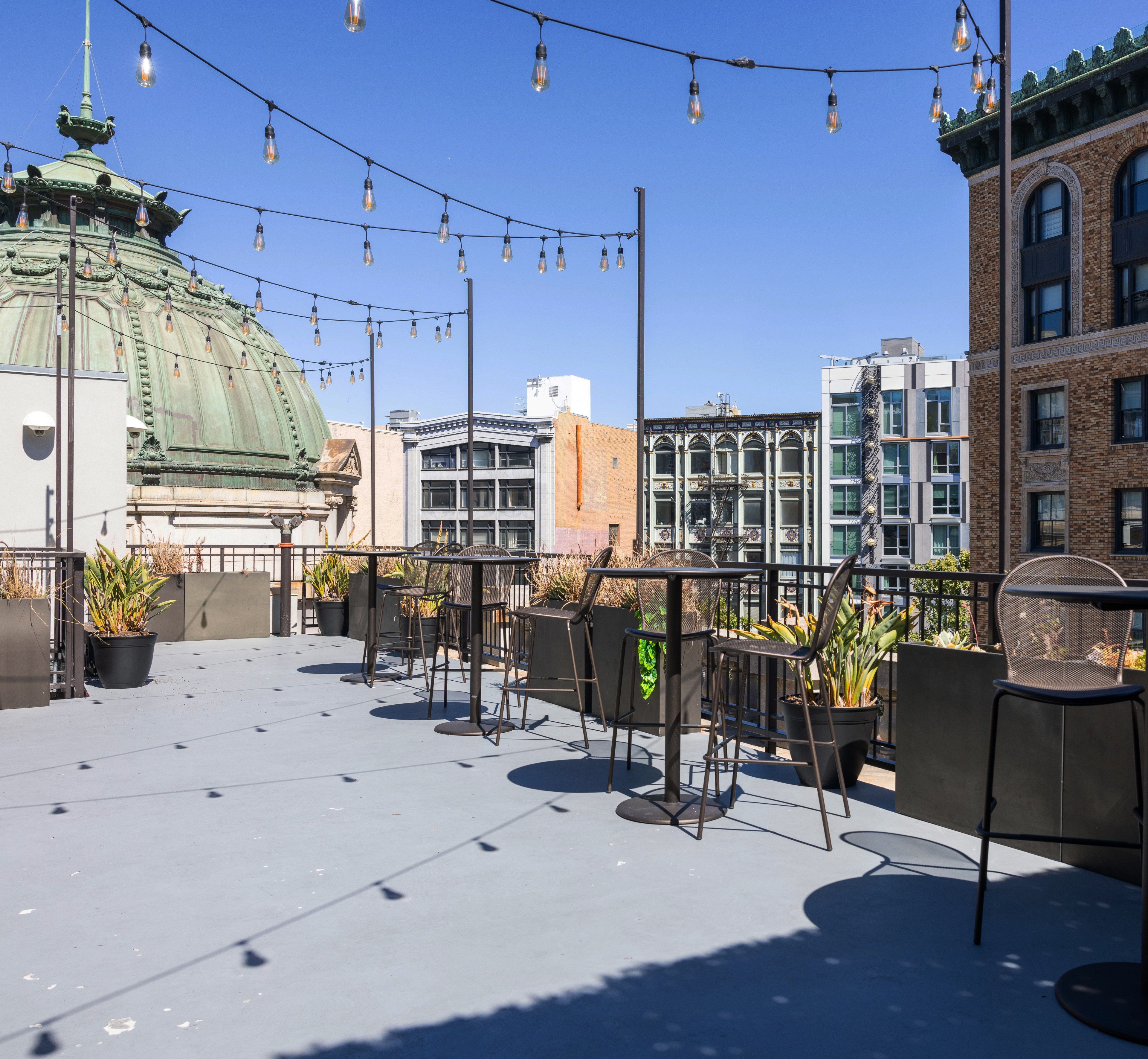 The image shows a rooftop patio with string lights, high tables, and barstools, surrounded by planters with greenery. In the background, there are urban buildings and a green domed structure.