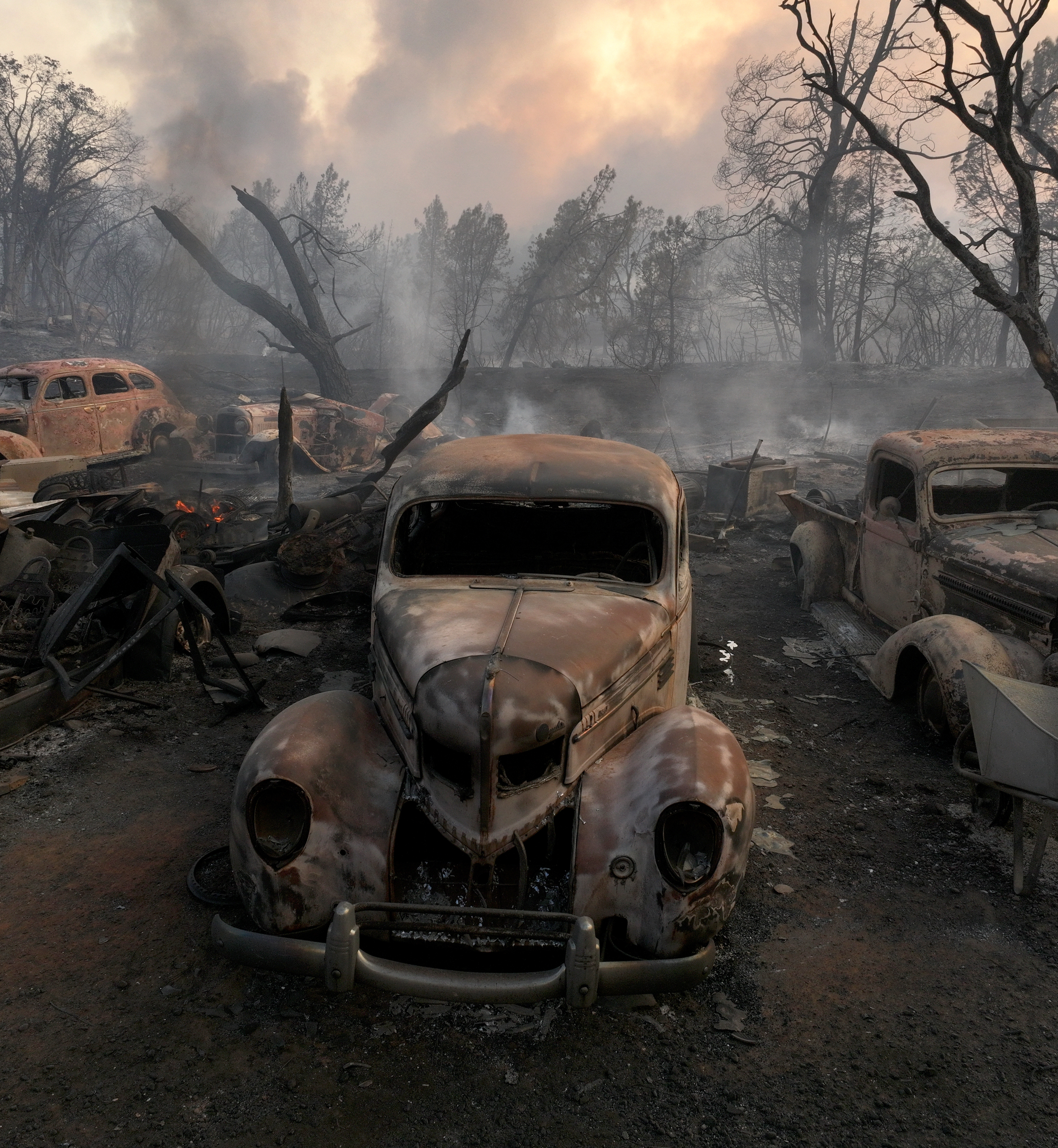 The image shows a desolate scene with burned, rusted old cars amid charred remains, smoke, and leafless trees; the aftermath of a devastating fire.