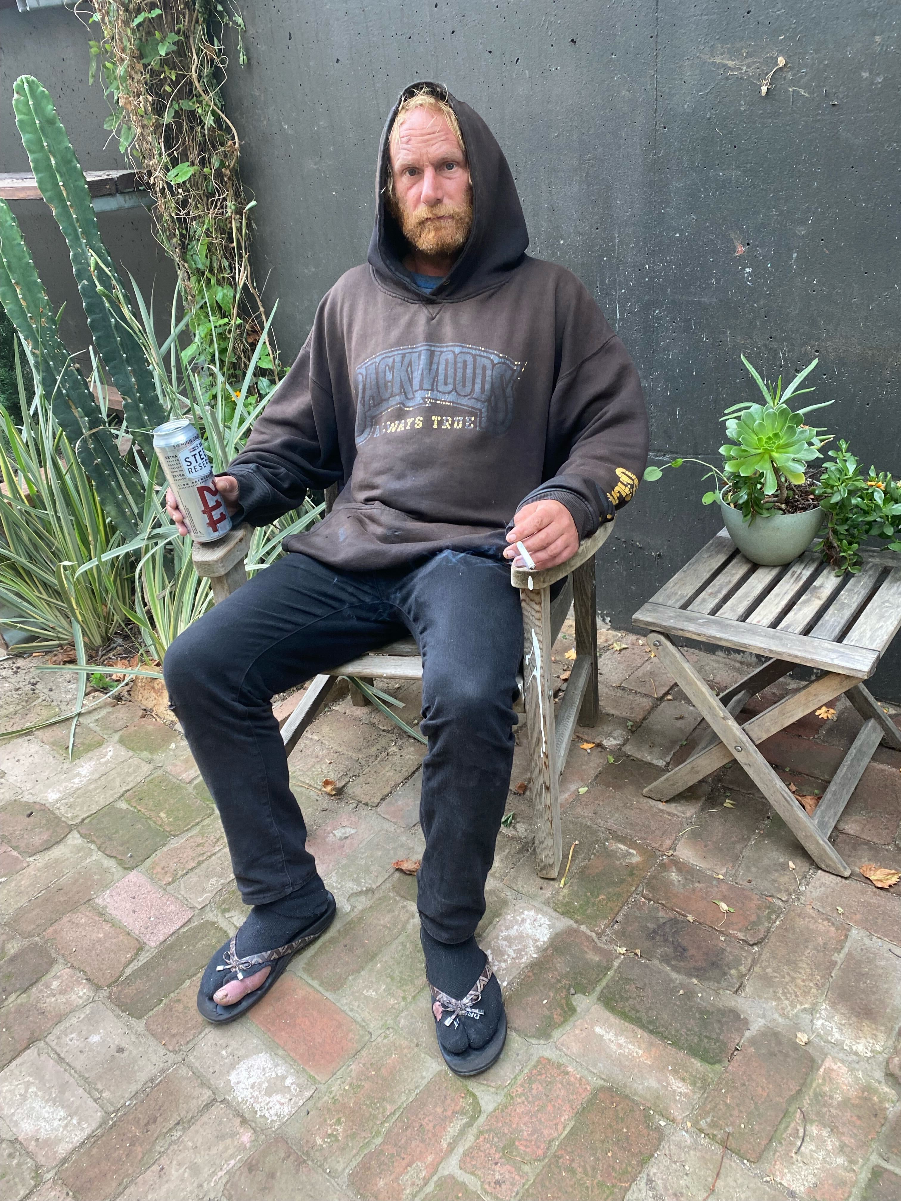 A man with a beard, wearing a brown hoodie, black pants, and flip-flops with socks, sits in a wooden chair outdoors holding a beer can and a cigarette. There's a cactus and potted plant beside him.