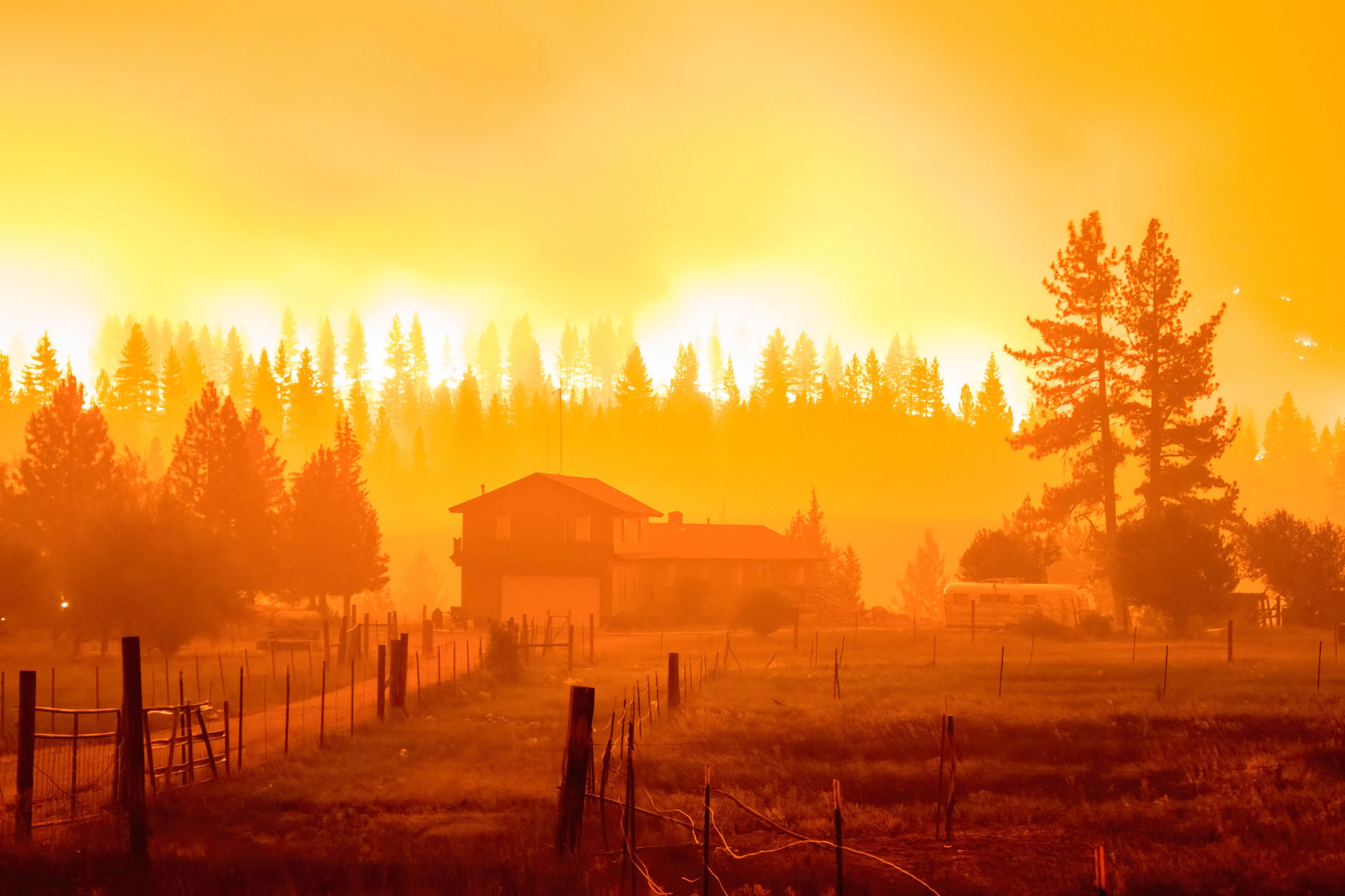 A house stands amidst a smoky field with a blazing forest fire in the background, casting an intense orange glow over the sky and surrounding trees.