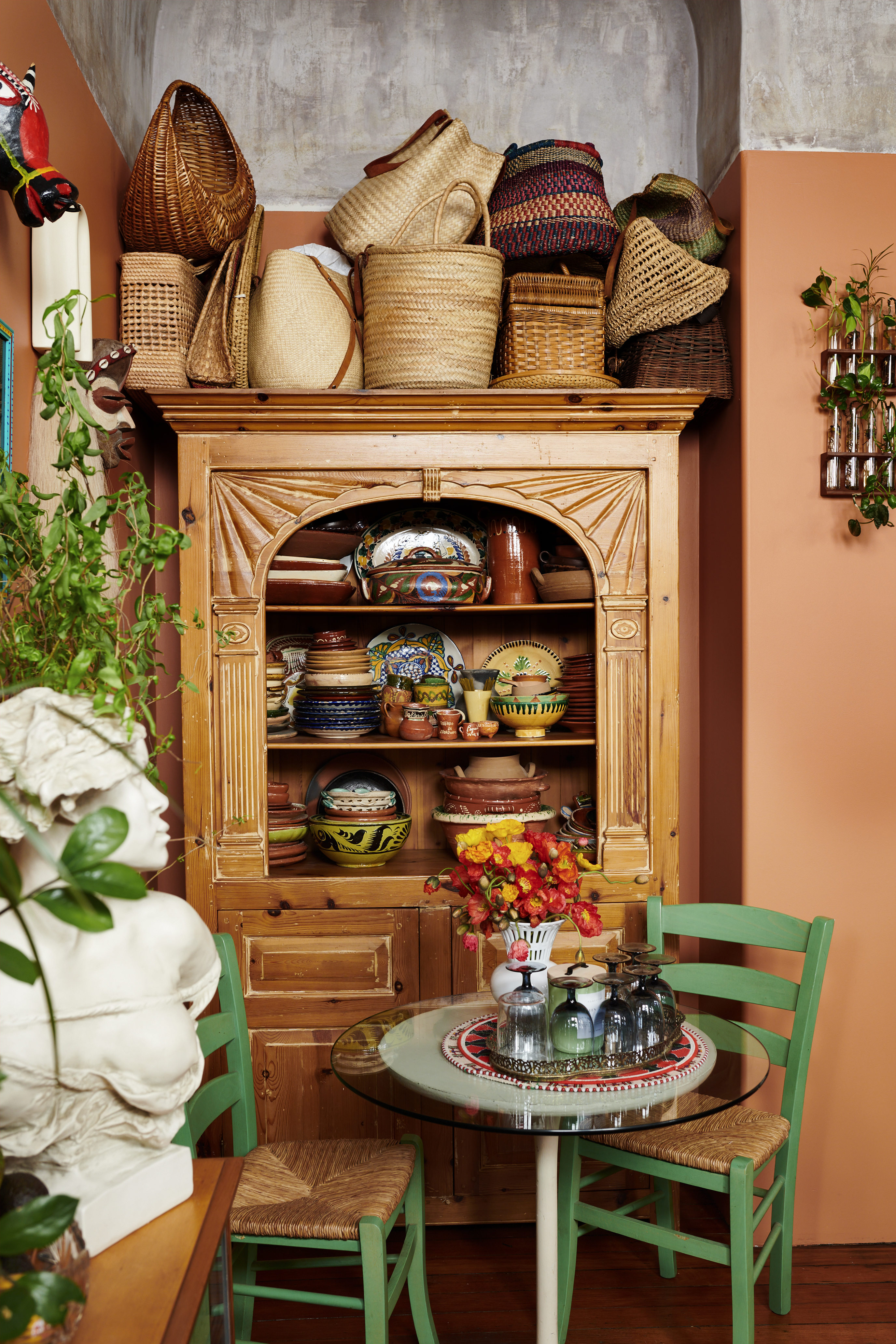 A wooden cabinet filled with colorful ceramics and tableware, topped with wicker baskets, stands next to a glass table with green chairs, adorned with flowers and glassware.