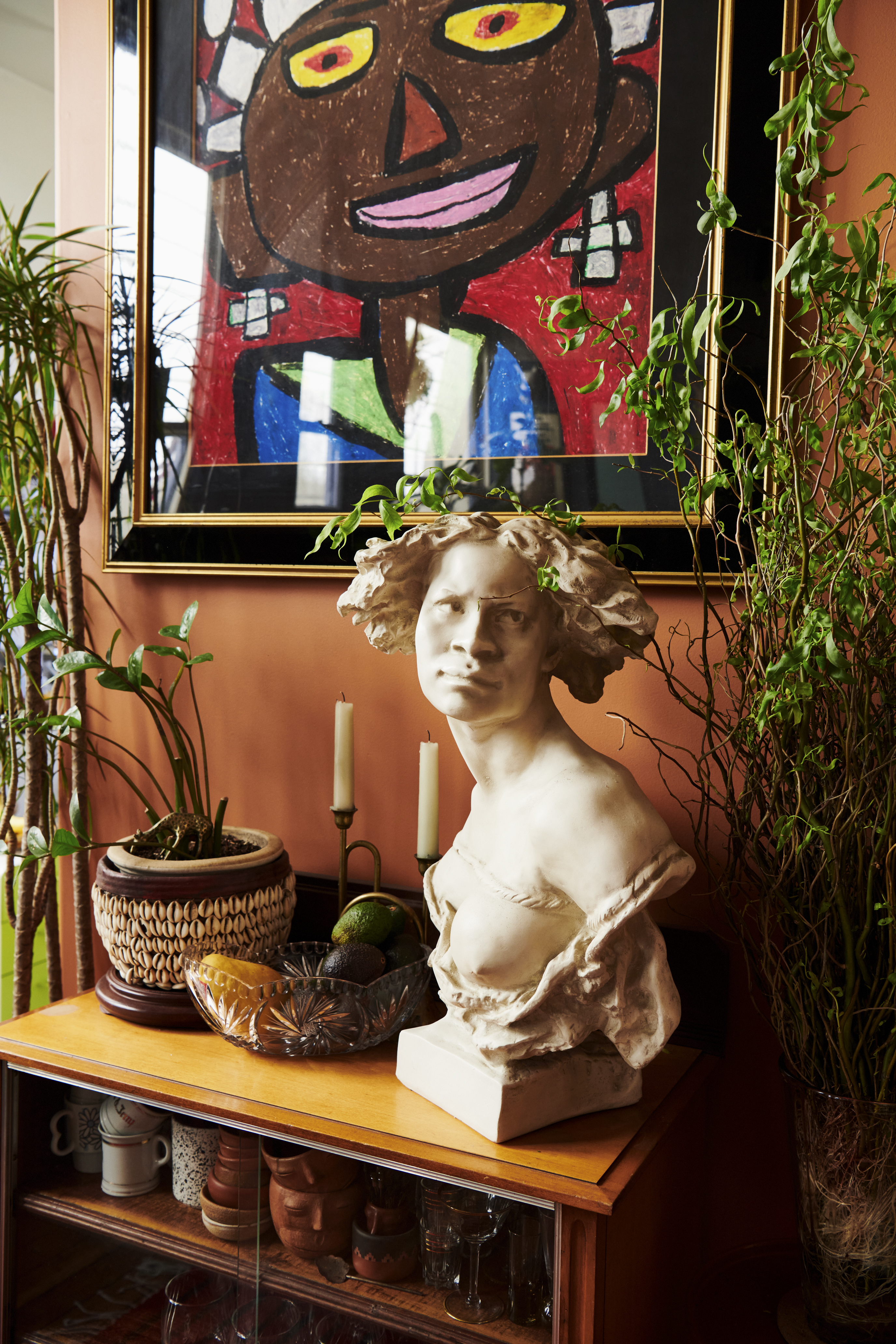 A white marble bust sits on a wooden cabinet topped with plants, fruit, and candles, beneath a vibrant framed painting of a smiling face.