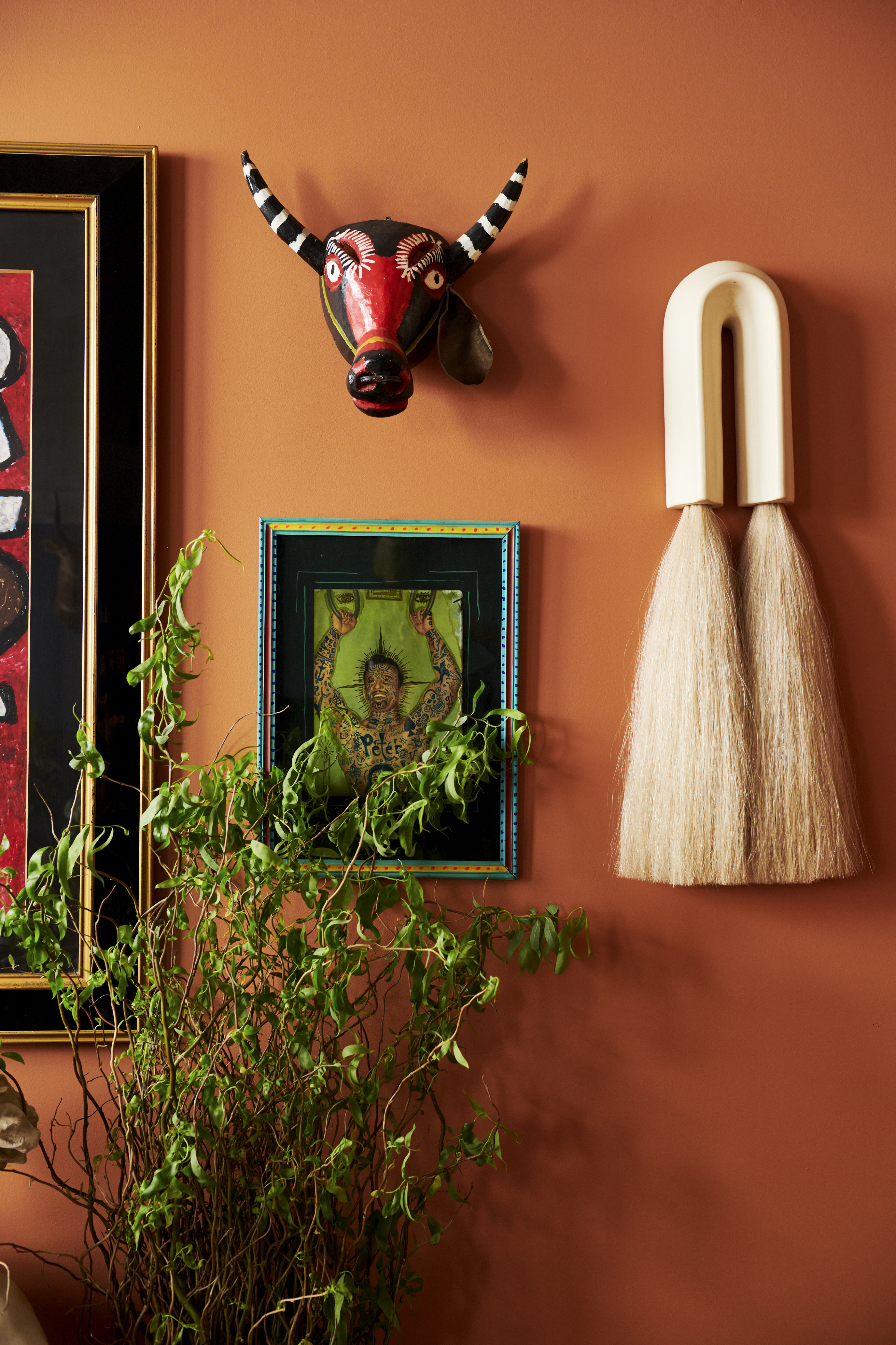 The image shows a warm orange wall adorned with a red and black bull mask, a framed artwork, and a decorative piece with blonde tassels. A lush green plant sits below.