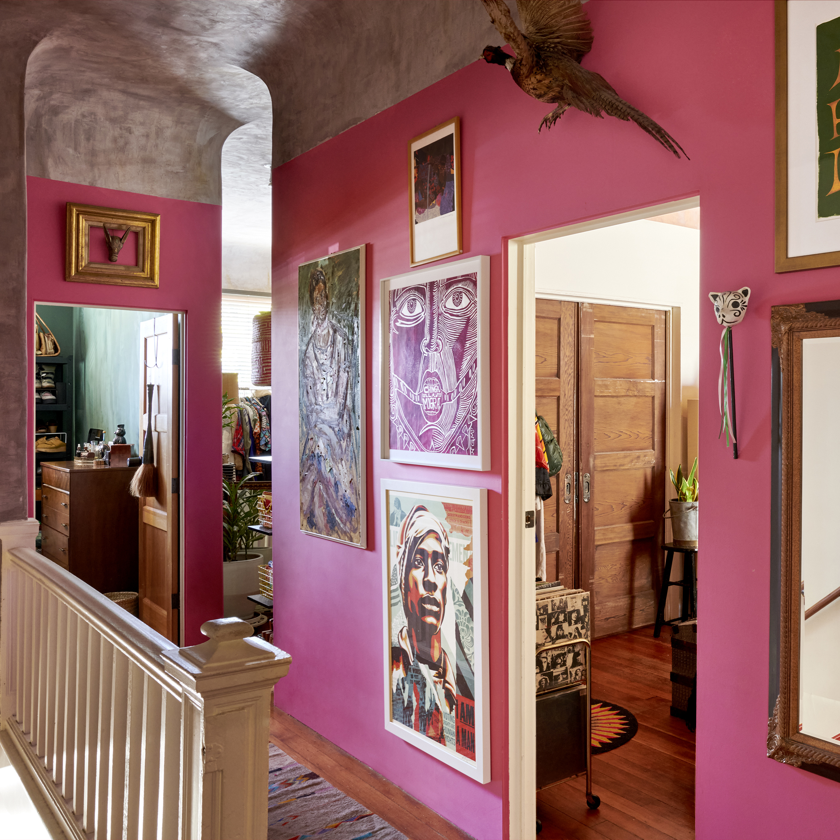 The image shows a vibrant hallway with pink walls adorned with various framed artworks, leading to rooms with wooden doors. A ceiling-high arch is visible on the left side.