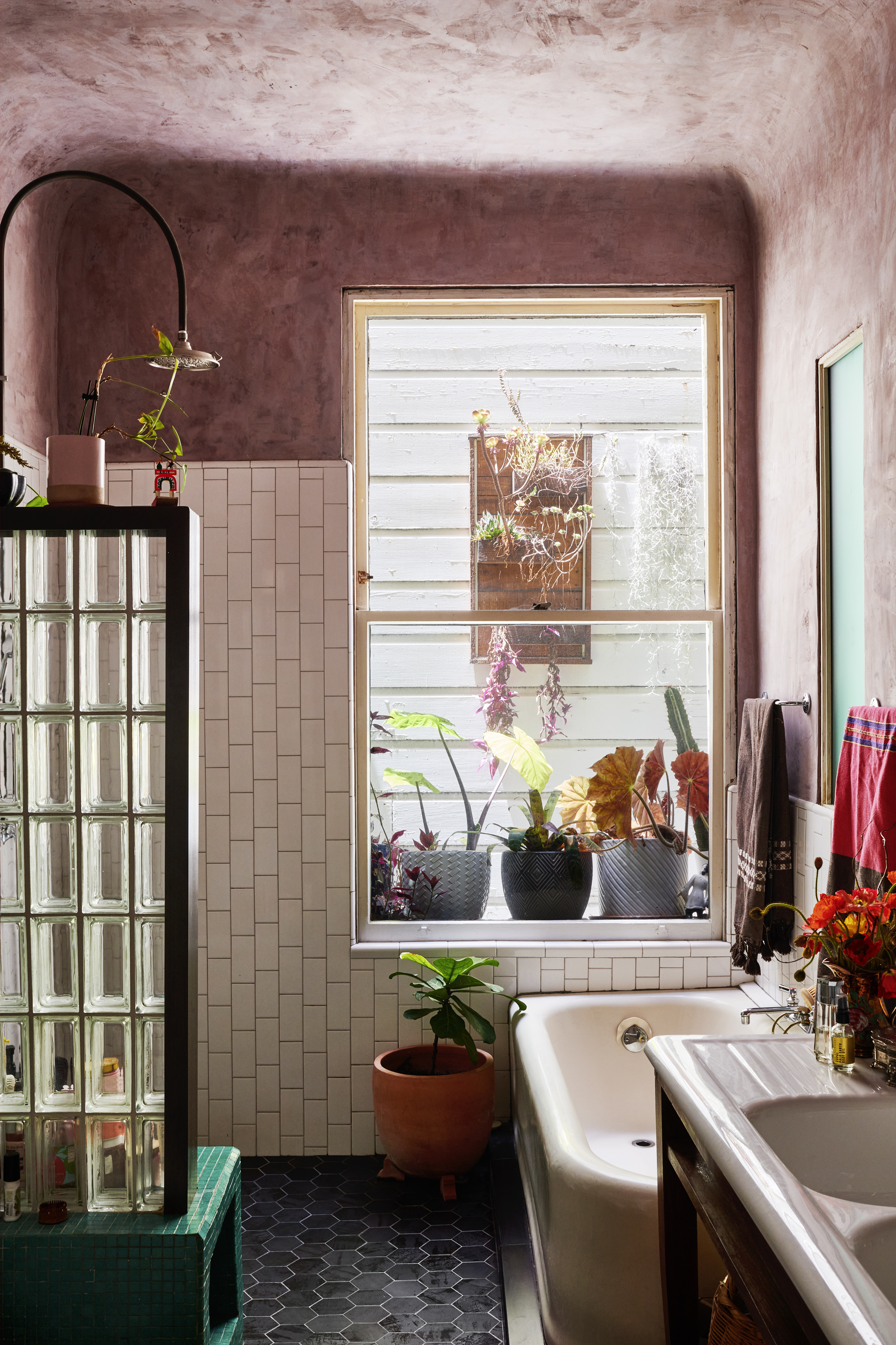 The bathroom features a pinkish ceiling, tiled walls, a glass block divider and a bathtub. A window showcases various potted plants and hanging decorations with colorful towels.