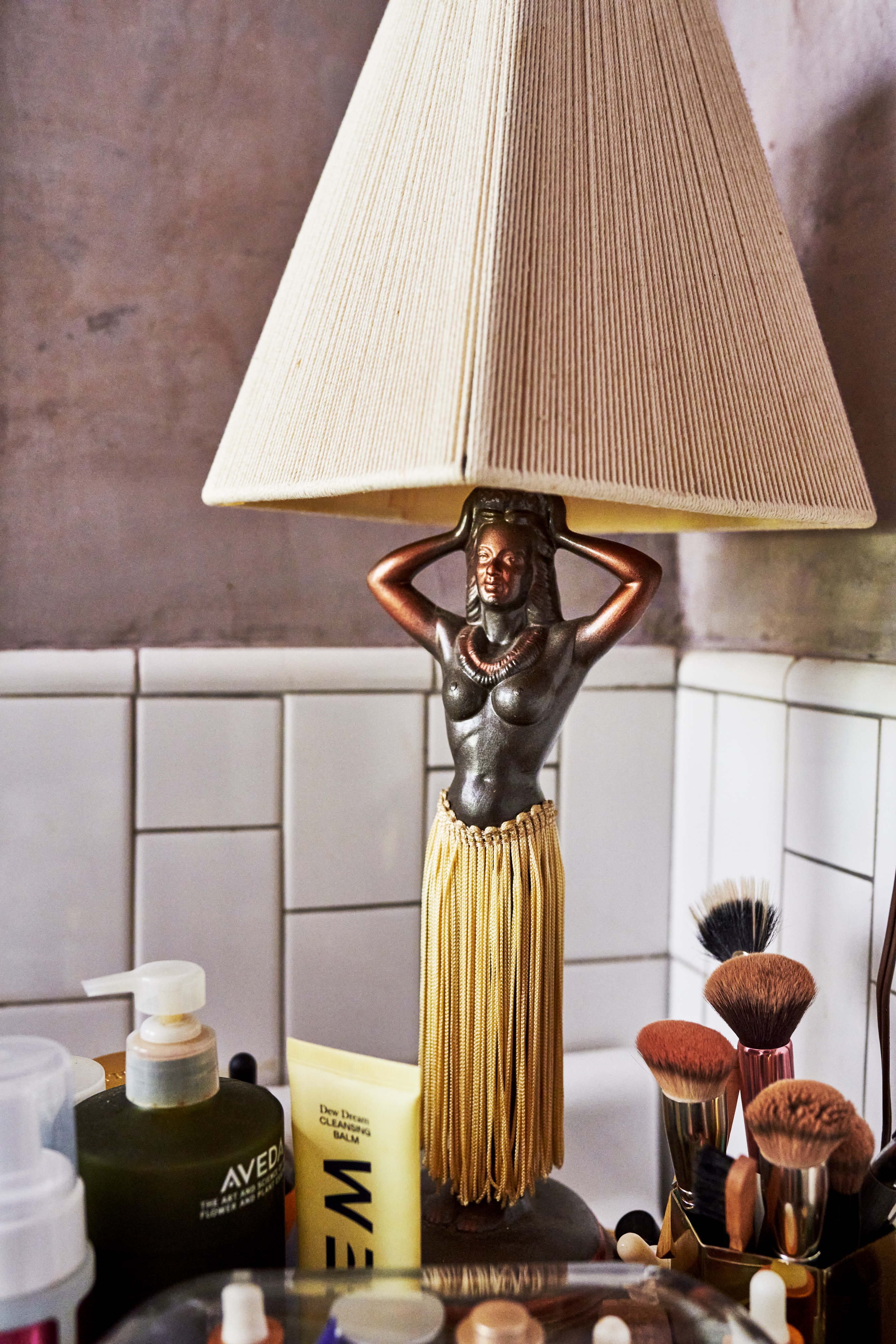 The image shows a decorative lamp with a woman figure base, fringed skirt, cream lampshade, surrounded by toiletries, makeup brushes, and beauty products. It has a tiled wall background.