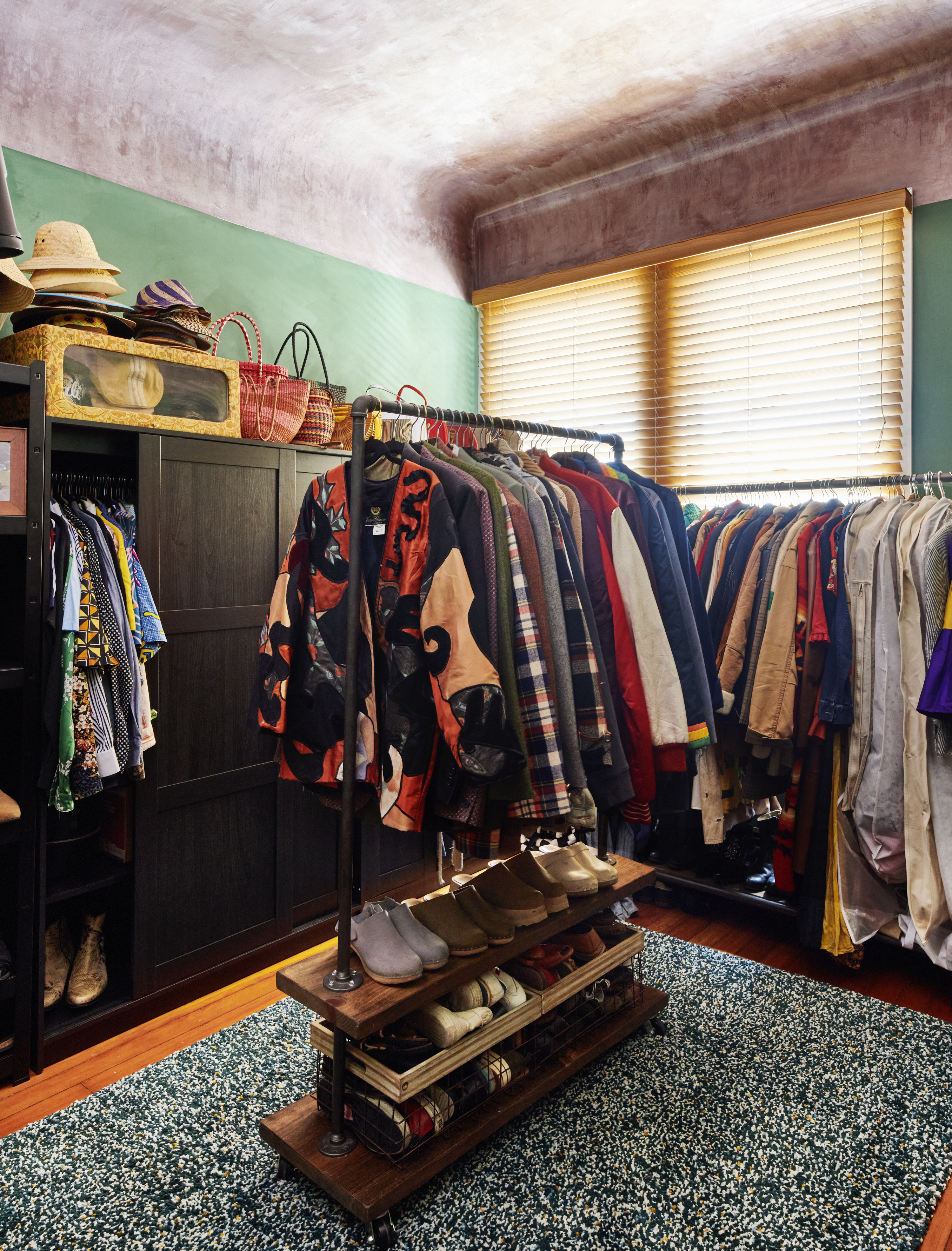 A cozy closet with hats on the top shelf, jackets and shirts hanging, shoes neatly arranged on shelves below, and a window with blinds brightening the space.