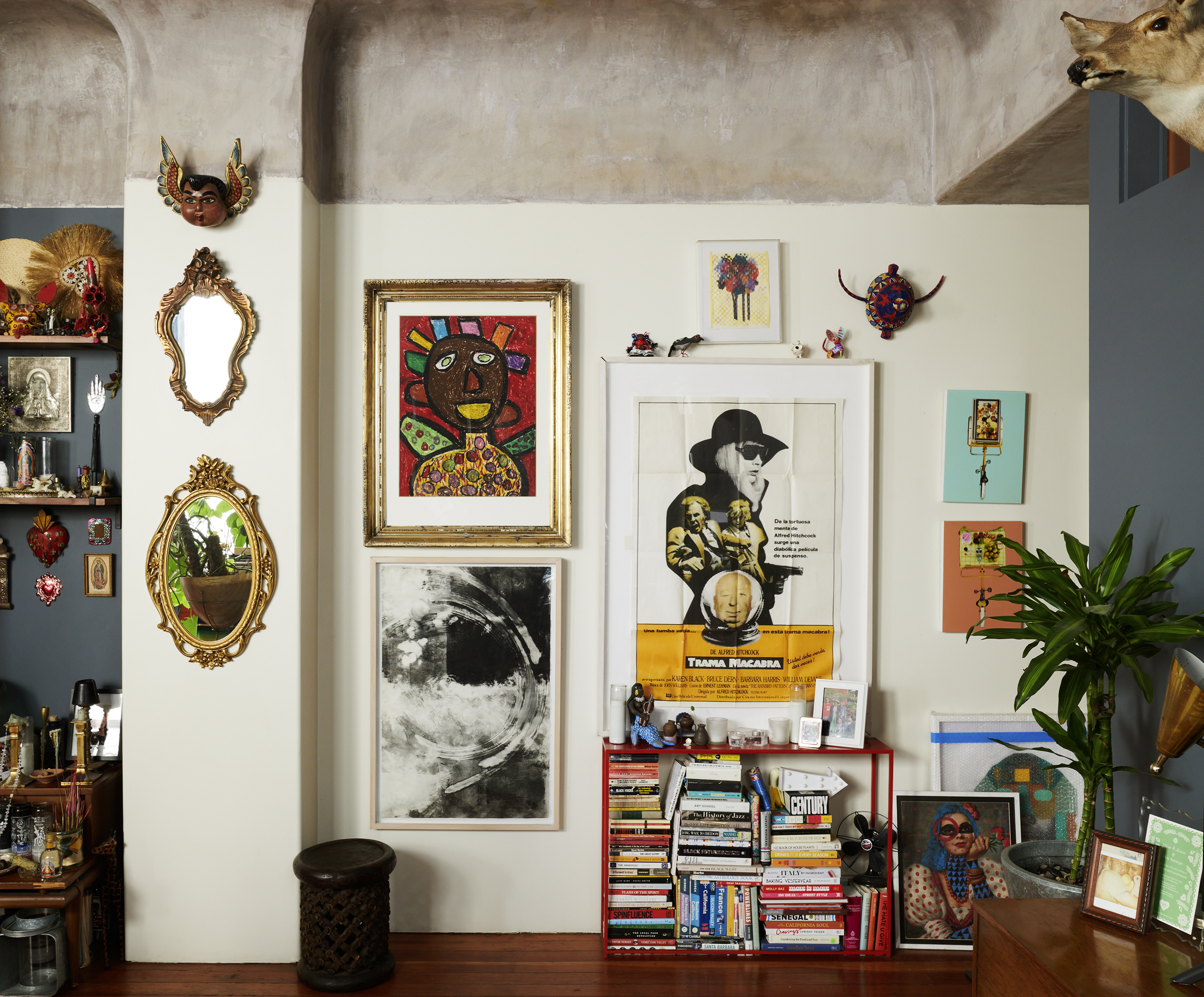 The image shows an eclectic living space with various framed artworks, a bookshelf filled with books, a plant, mirrors, a deer head mount, and other decorative items and trinkets.