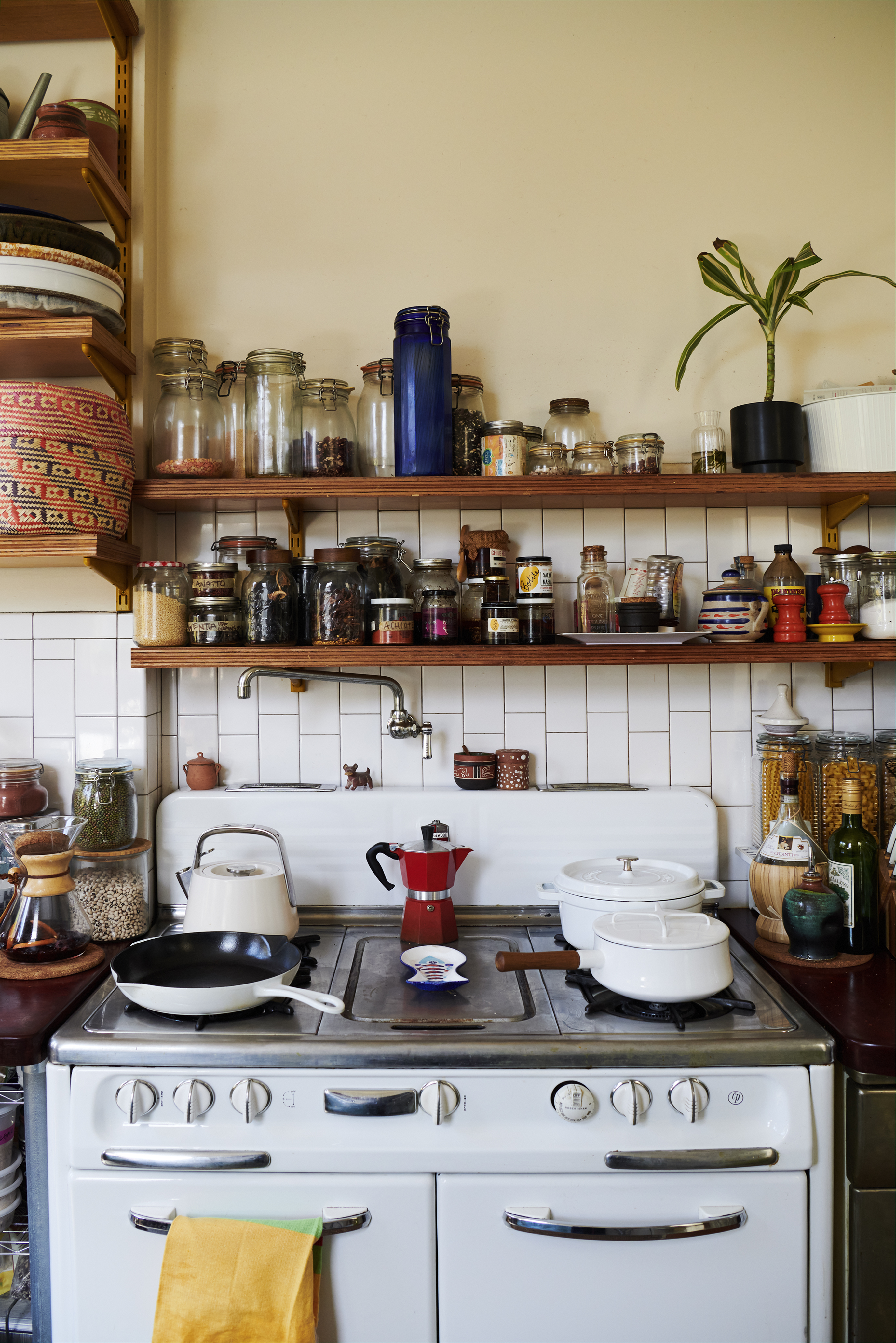 This image shows a cluttered kitchen with jars of spices on shelves, a white stove with various cooking pots and a red coffee maker, and a few plants.