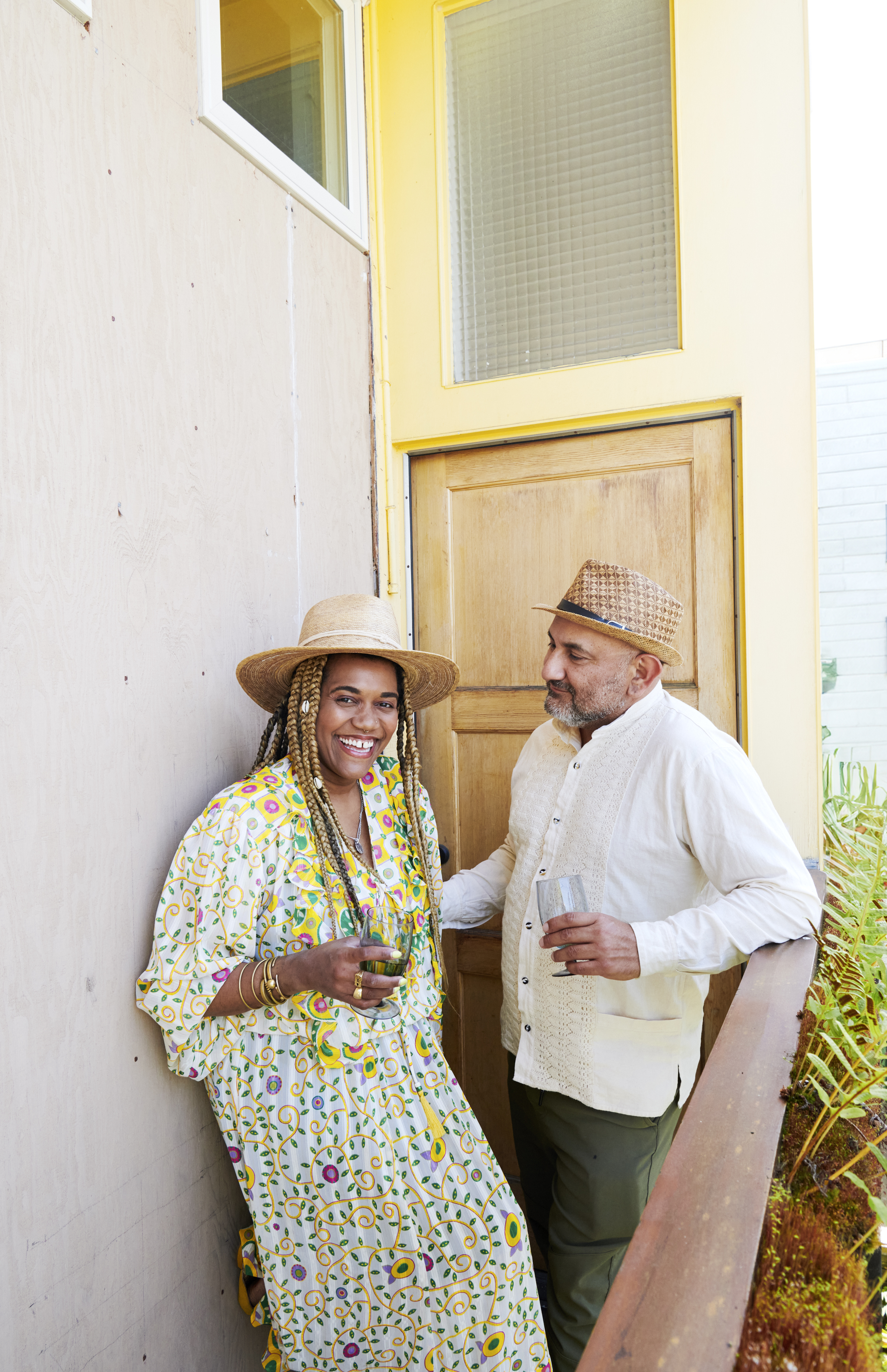 Two people stand outside a door, both holding drinks. One wears a colorful dress and straw hat, while the other is in a white shirt and hat, smiling and chatting.