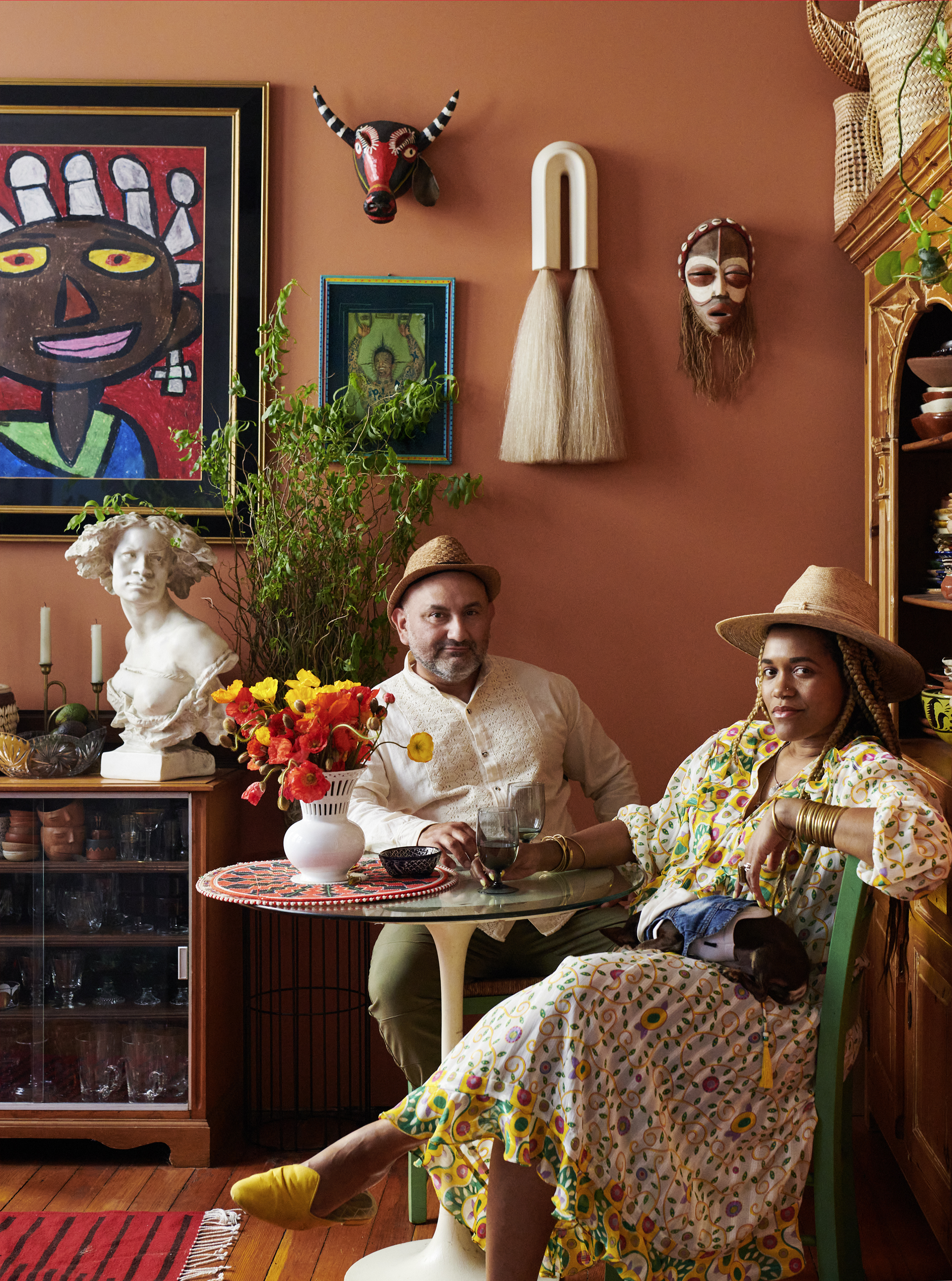 Two individuals in hats sit at a round table with flowers, in a colorful room adorned with art, masks, plants, and sculptures, creating an eclectic ambiance.