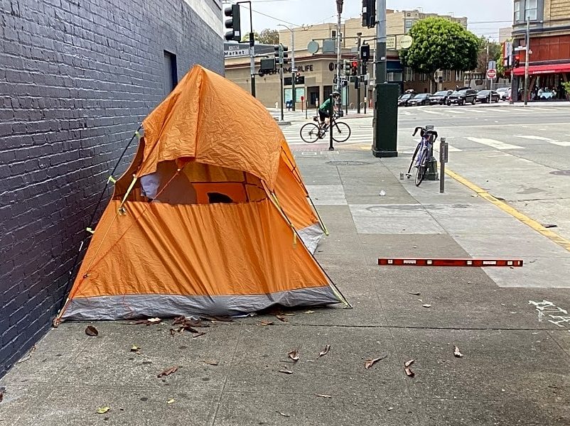 An orange tent is set up on a sidewalk against a brick wall in an urban area, with a bicycle and street intersection visible in the background.