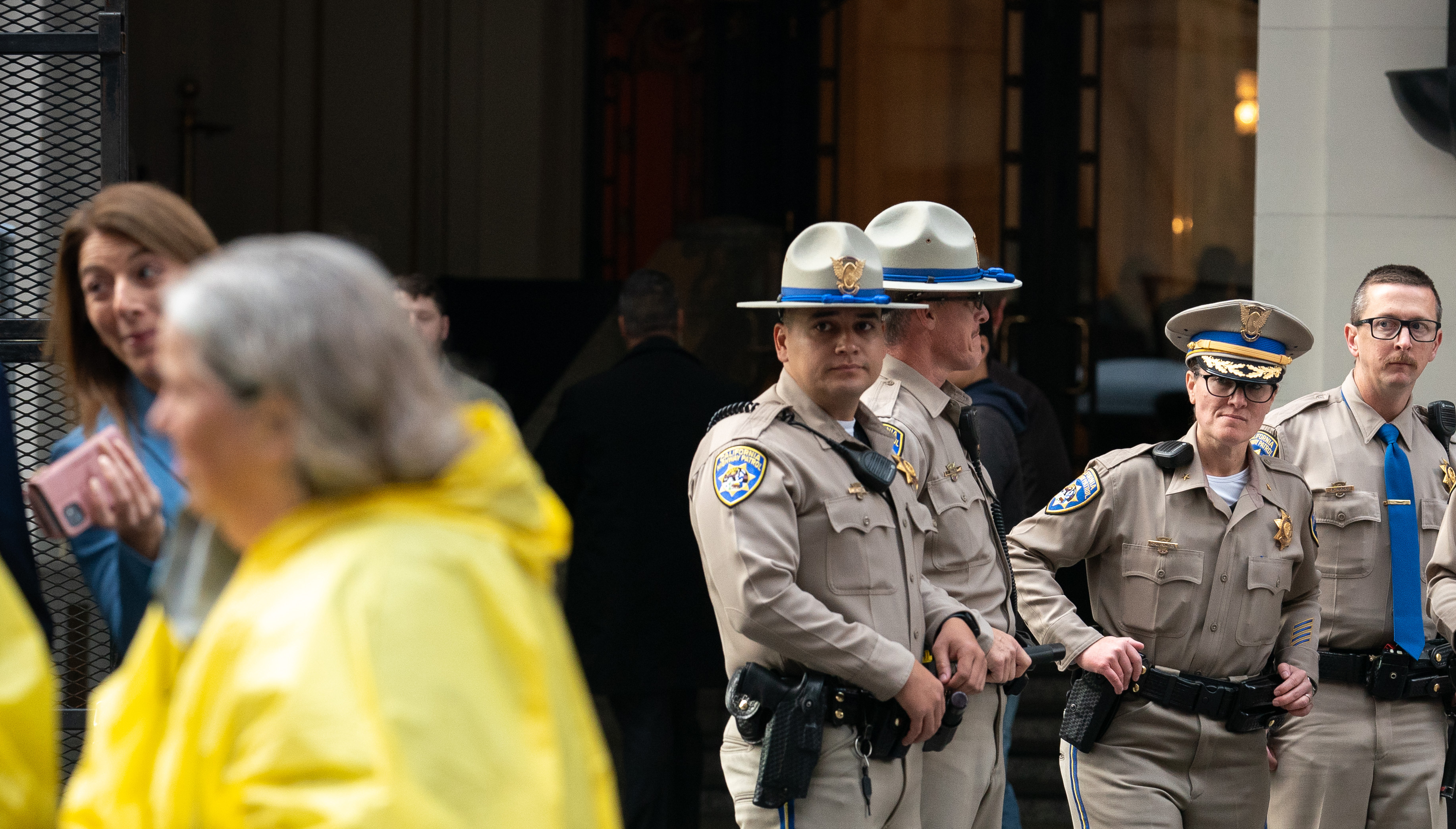 Several uniformed police officers with badges stand in a line; one officer looks at the camera. People in yellow raincoats and a woman with a pink phone walk by.