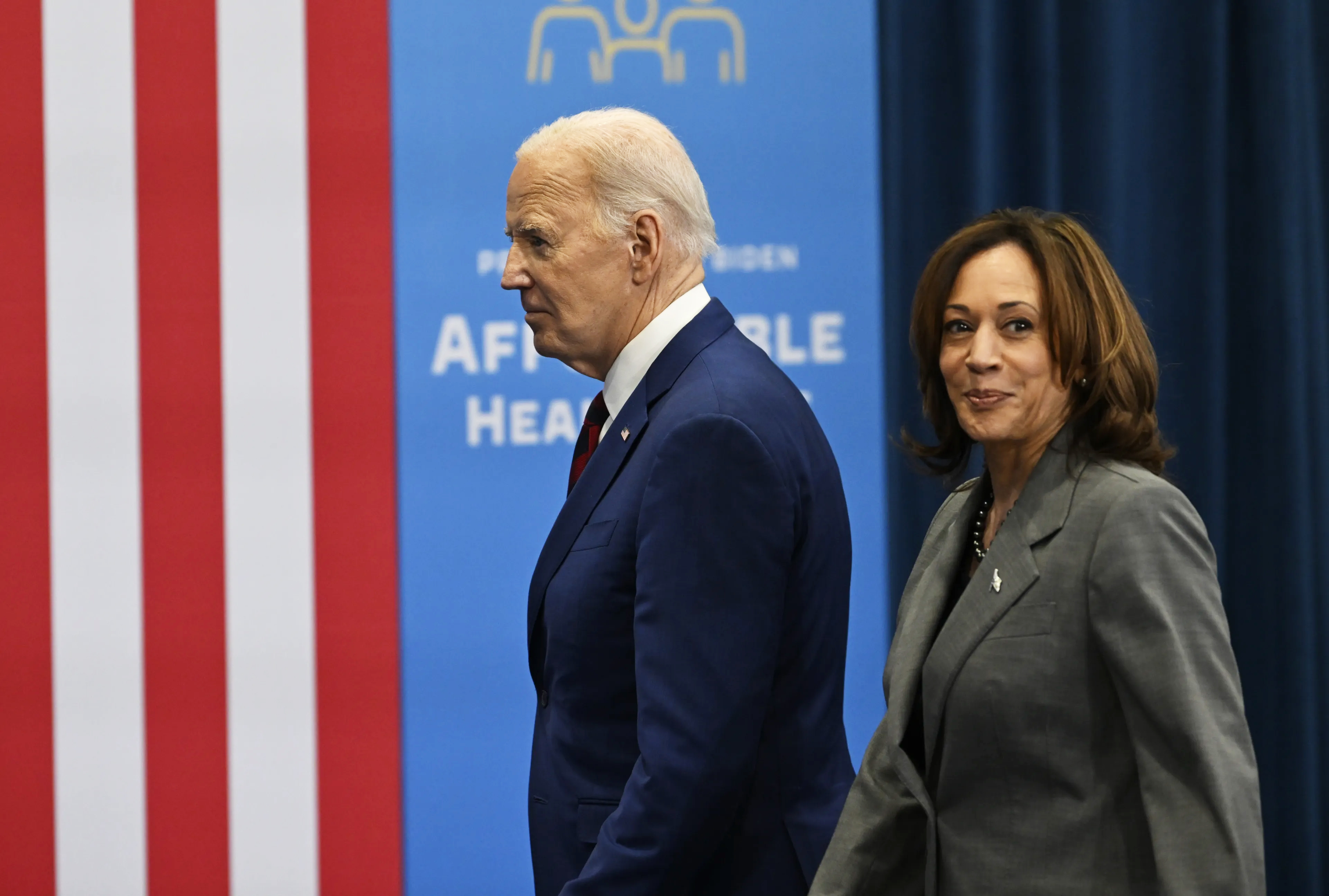 A man in a blue suit and a woman in a gray suit walk in front of a backdrop with the U.S. flag and text promoting affordable healthcare.