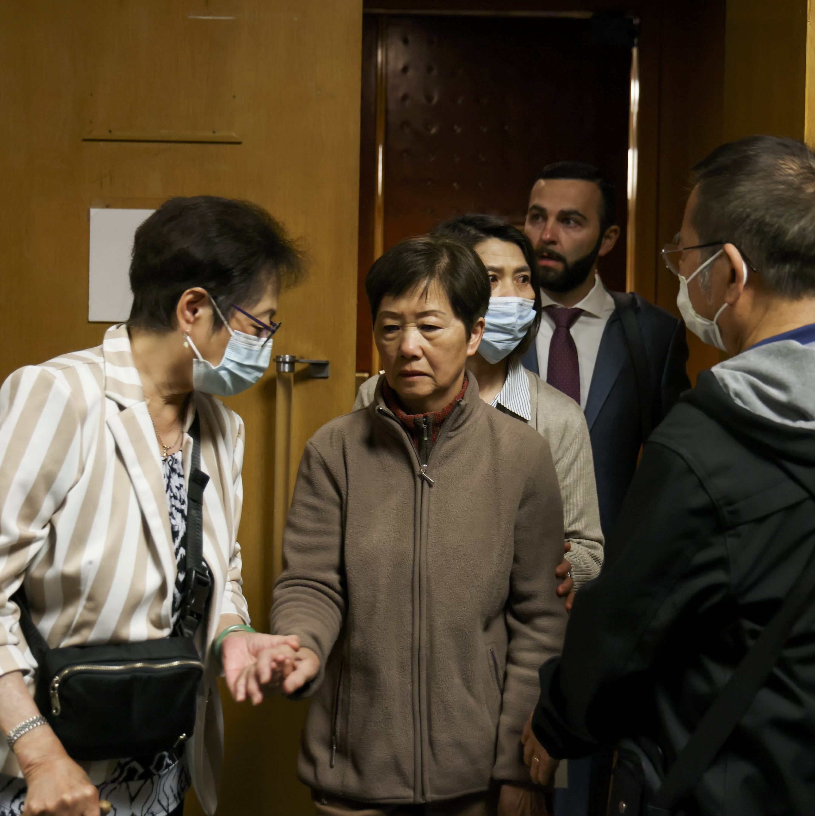 A group of five people are standing close together indoors. Two women in front wear surgical masks, while others look serious. One woman is extending her hand forward.