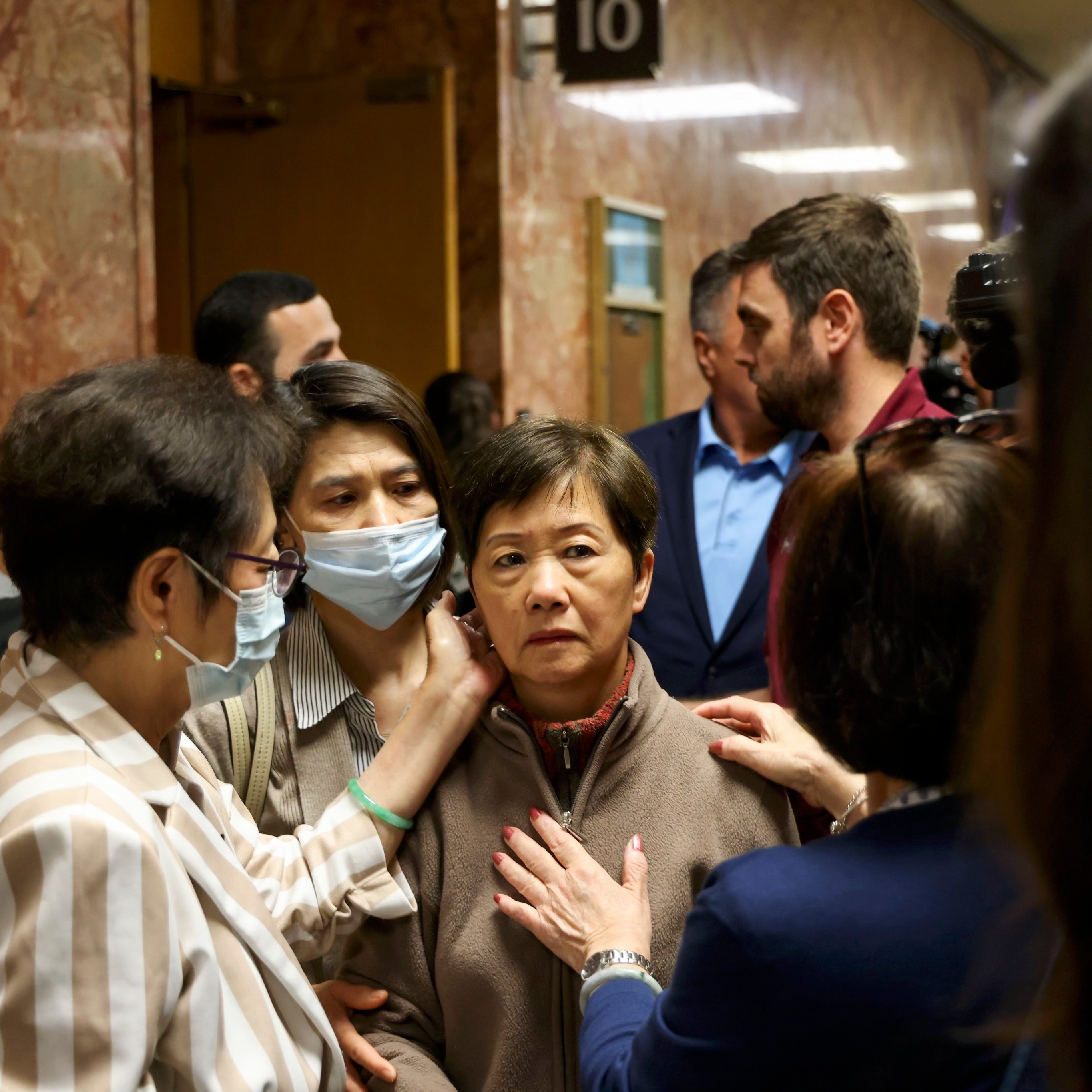 A woman wearing a mask that covers her mouth and nose caresses the face of a woman in a crowded hallway outside of a courtroom.