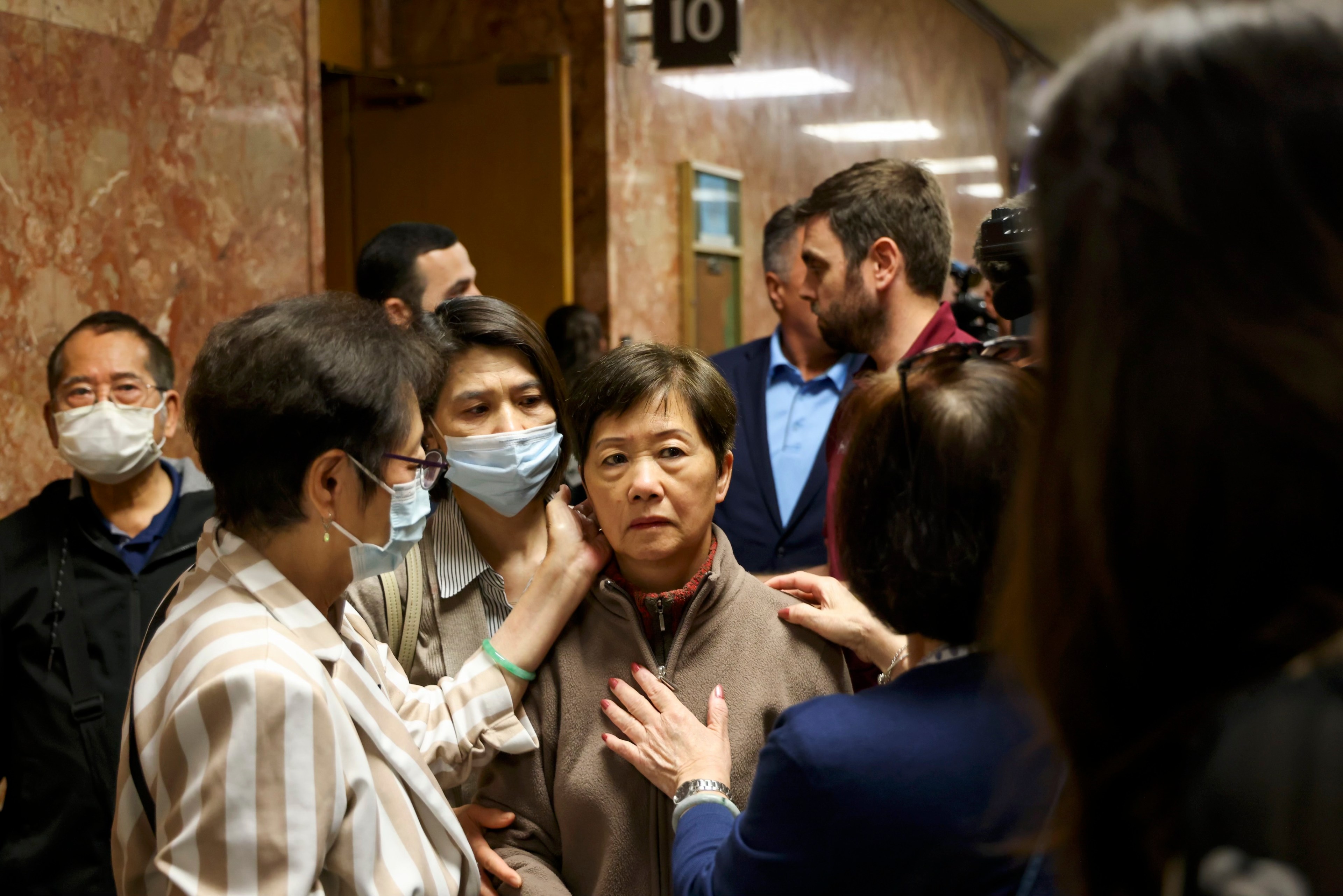 A woman wearing a mask that covers her mouth and nose caresses the face of a woman in a crowded hallway outside of a courtroom.