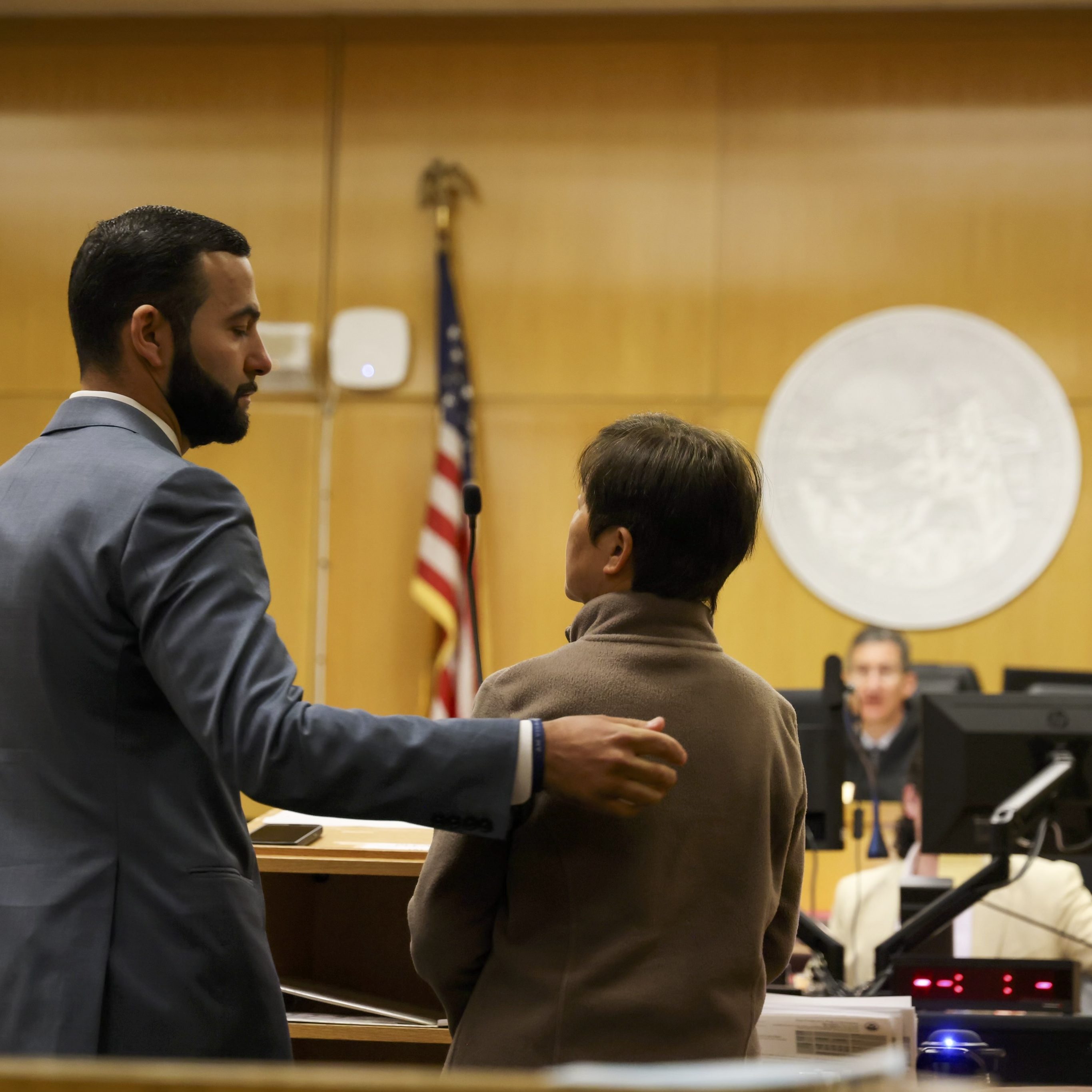 A man in a suit places his hand on a woman's shoulder in a courtroom, with an American flag and a large emblem in the background. Two people, likely judges or clerks, sit at a desk.