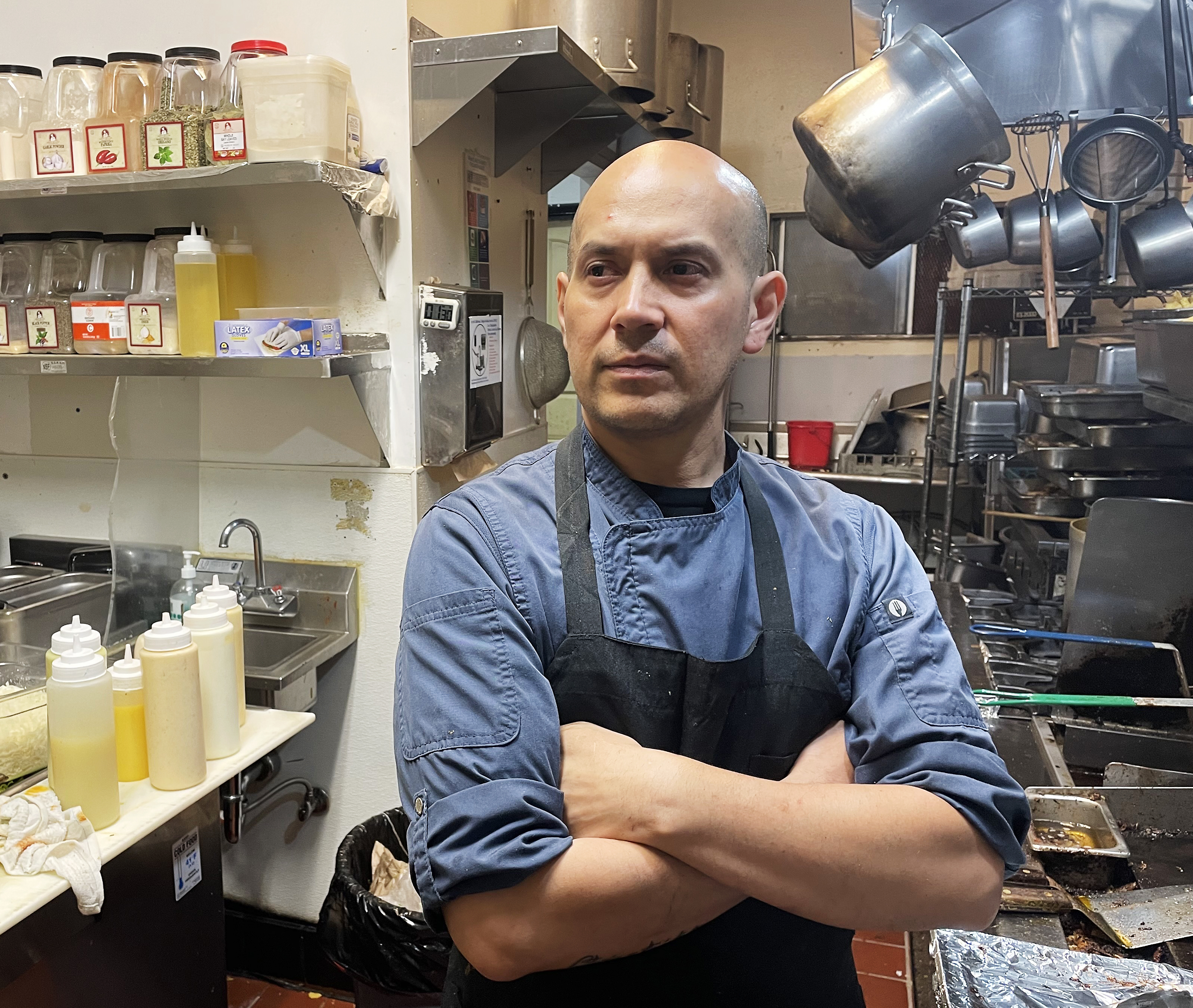 A chef with a shaved head stands in a commercial kitchen with arms crossed, dressed in a blue uniform and black apron. Shelves with spices and oils are on the left.