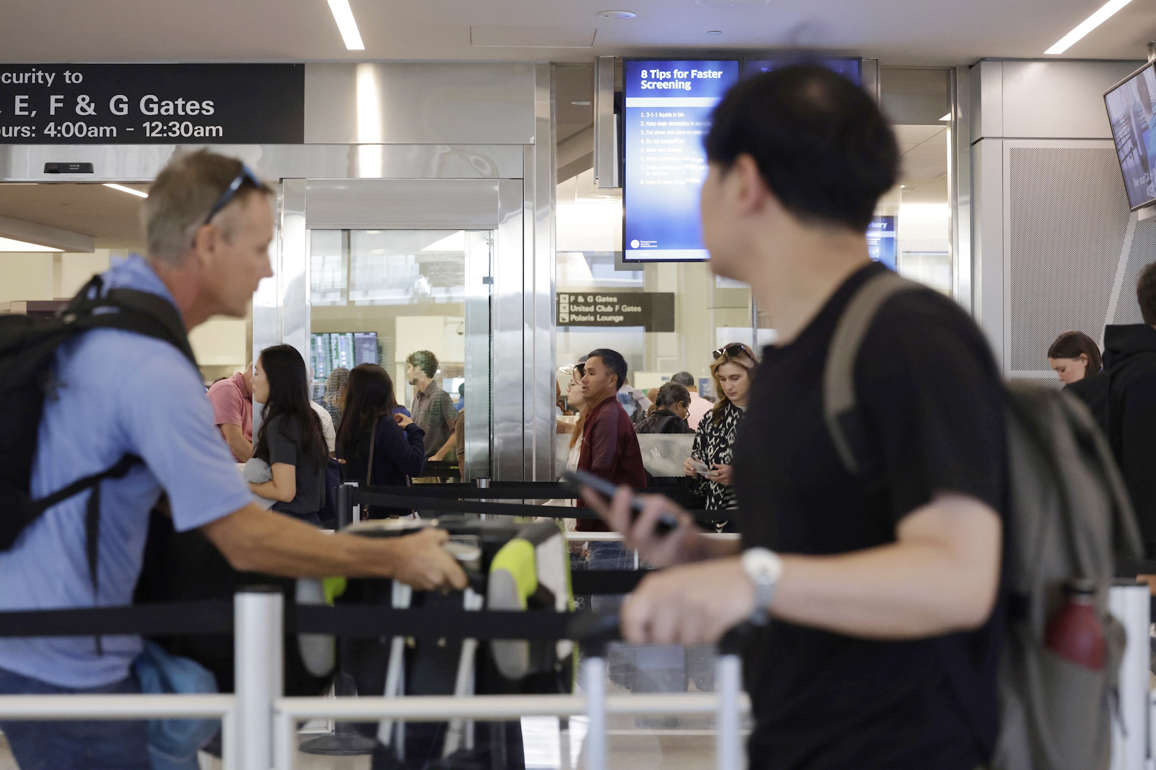 People are waiting in line at an airport security checkpoint, with signs and monitors visible overhead. Two men with backpacks are in the foreground.