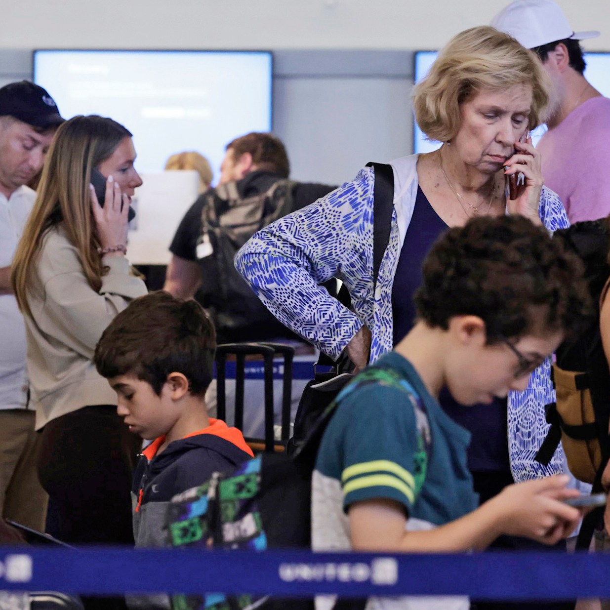 Several people, including children, stand in line at an airport, looking at their phones or talking. They appear focused and some show concern, with luggage nearby.