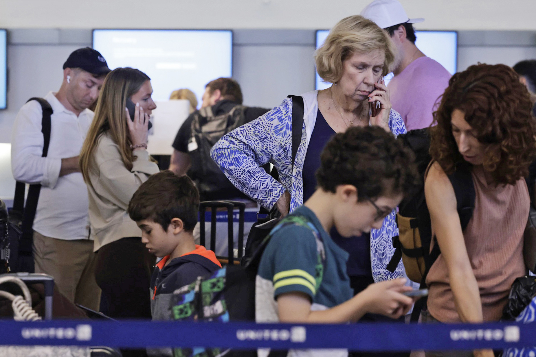 Several people, including children, stand in line at an airport, looking at their phones or talking. They appear focused and some show concern, with luggage nearby.