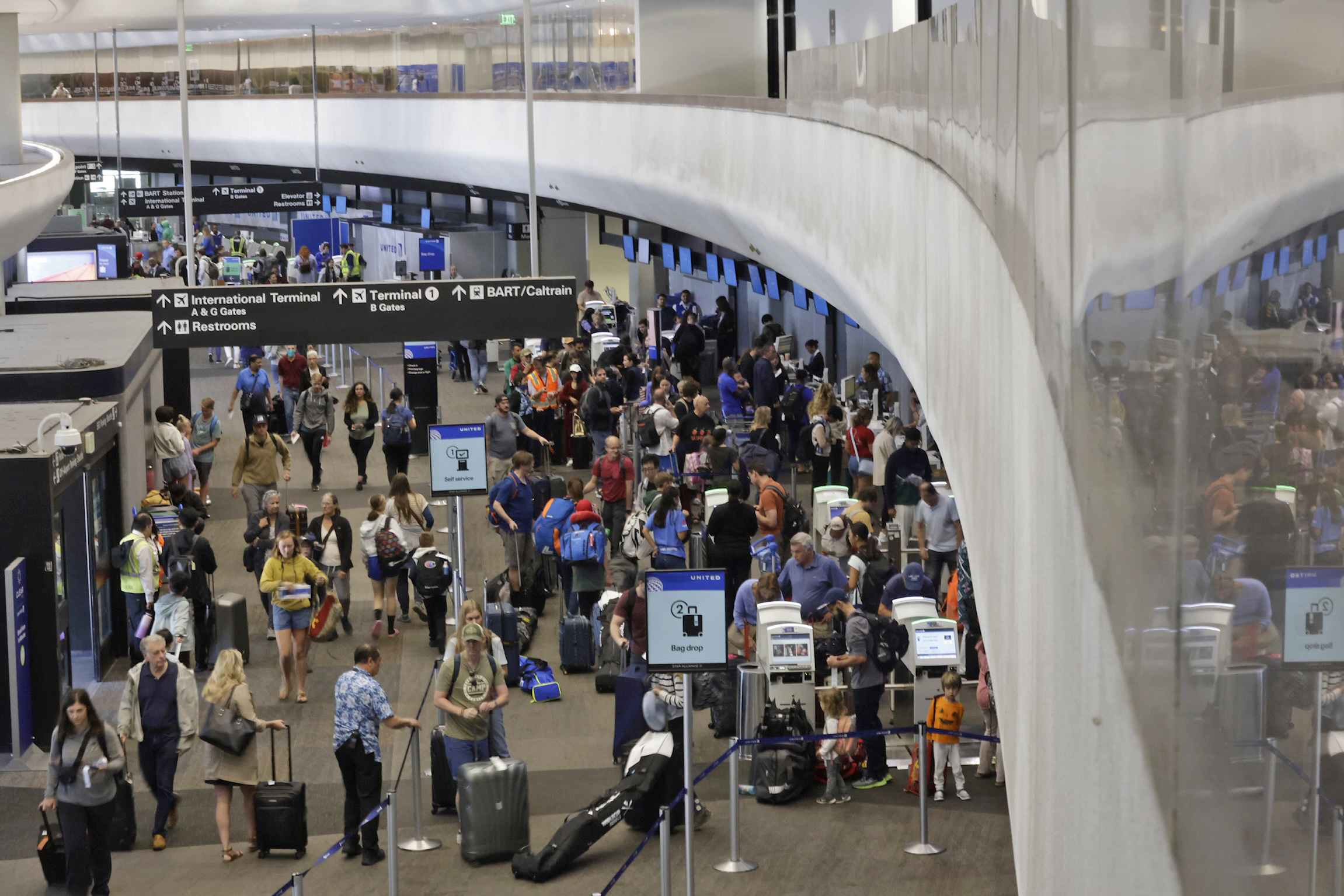 The image shows a busy airport terminal with many people walking around, checking in, and waiting in line. There are overhead signs directing travelers to various gates, terminals, and restrooms.