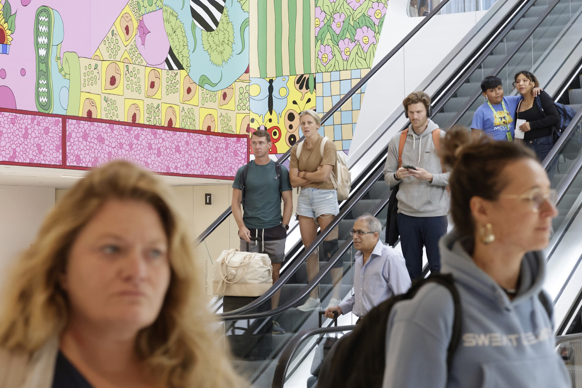 People move up and down escalators in a brightly decorated building with colorful murals. Some individuals look focused while others appear casual and relaxed.