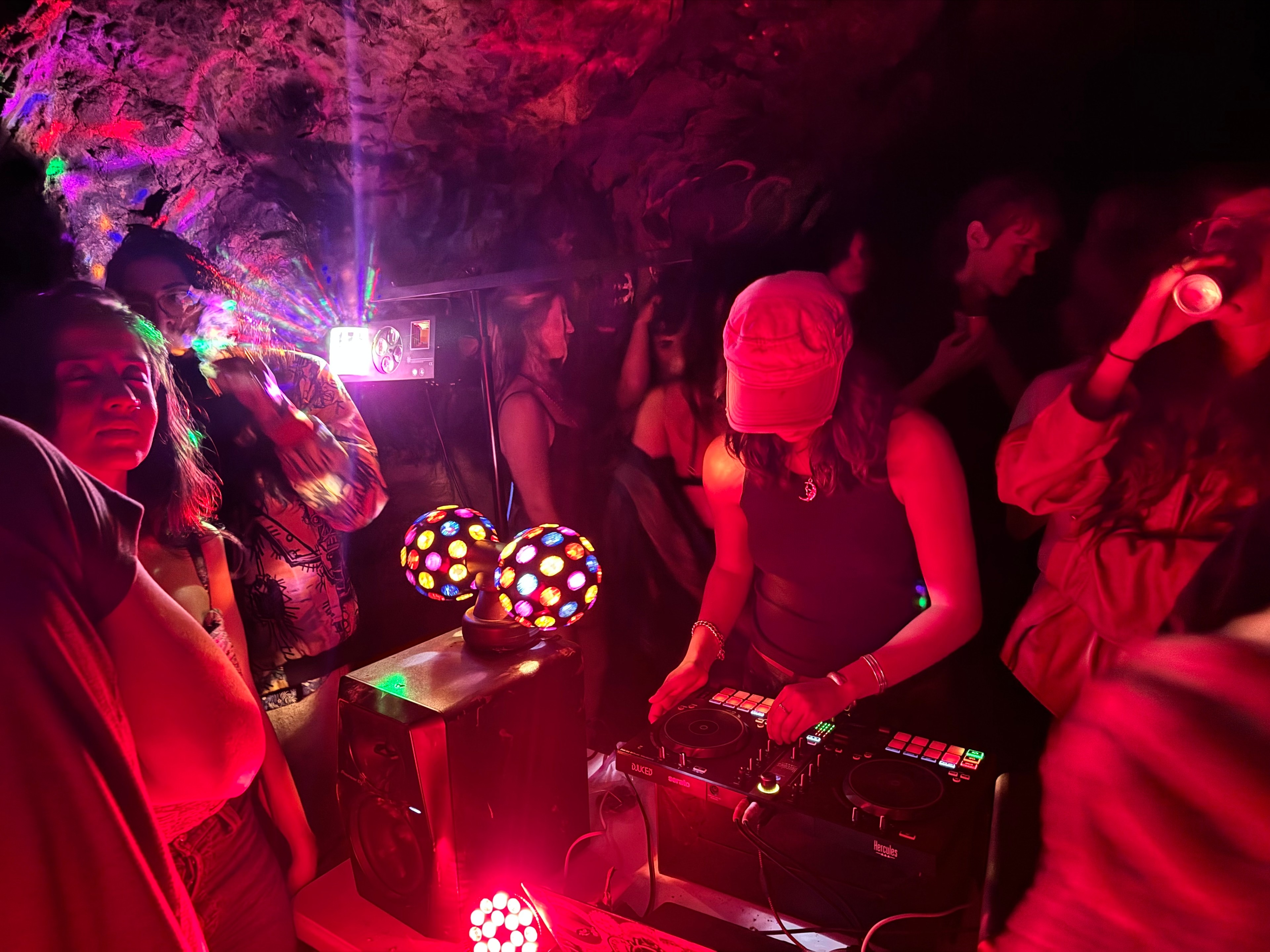 People gathered in a dimly lit, colorful underground party, with vibrant lights, a DJ wearing a cap playing music, and attendees dancing and socializing around.