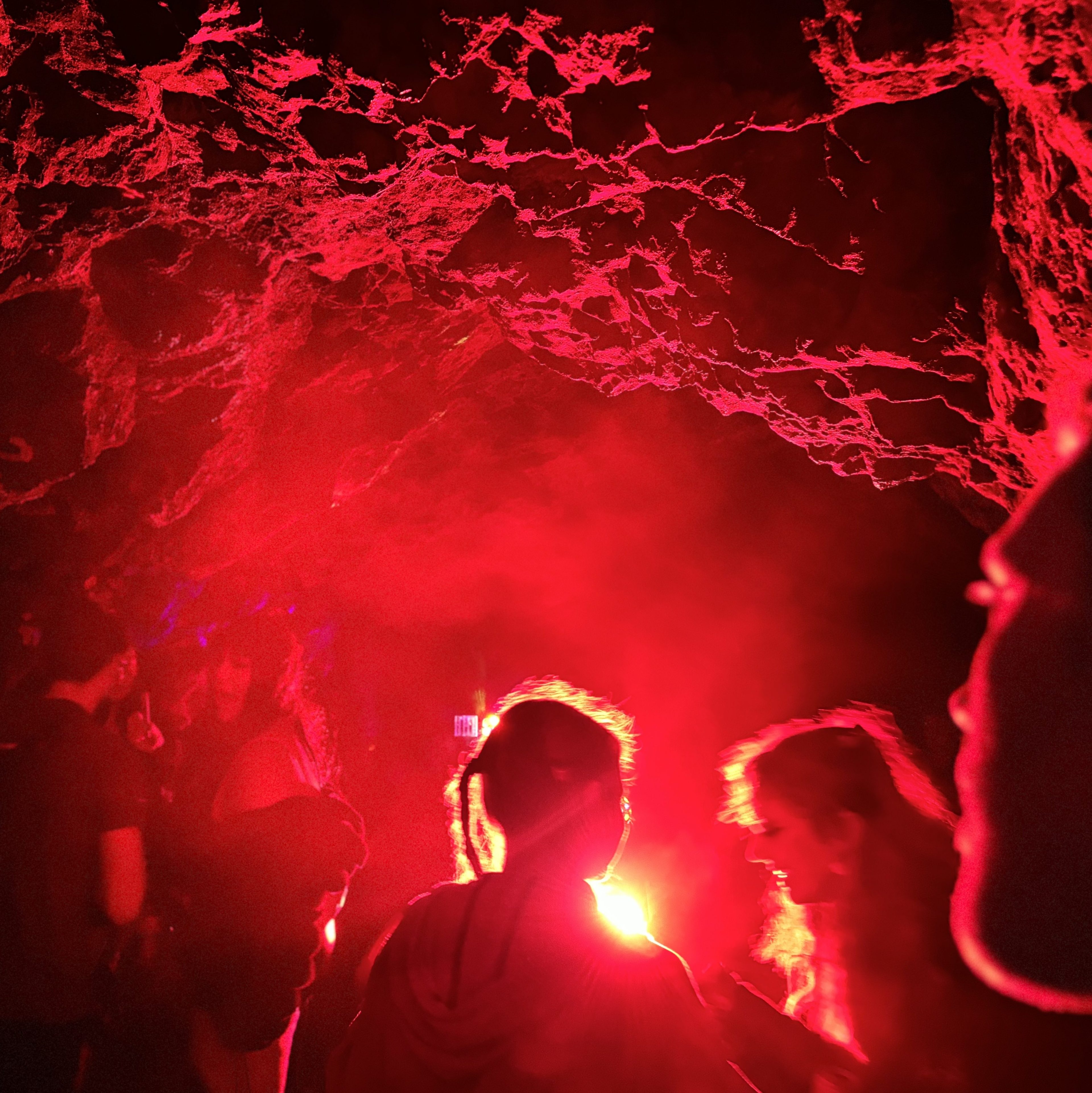 A group of people stands in a cave-like setting, illuminated by intense, red light. The rocky ceiling is visible, while the individuals appear engaged in conversation.