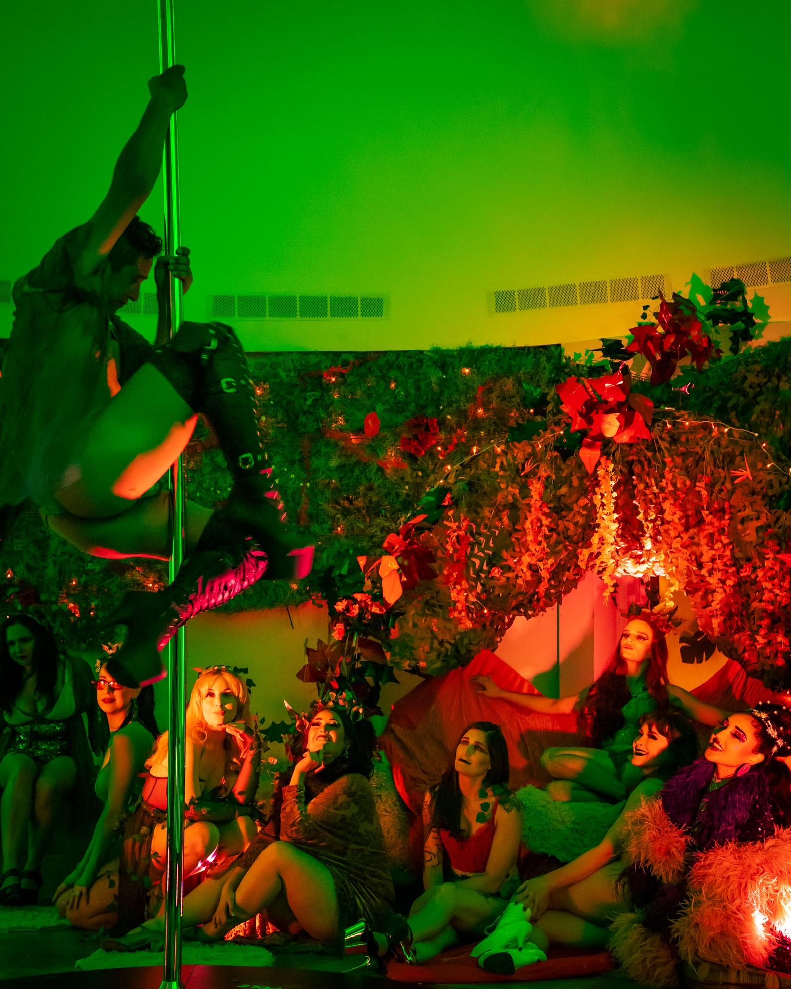 A performer in provocative attire is pole dancing under green lights, while a group of women in themed costumes eagerly watch from a lush, decorated background.