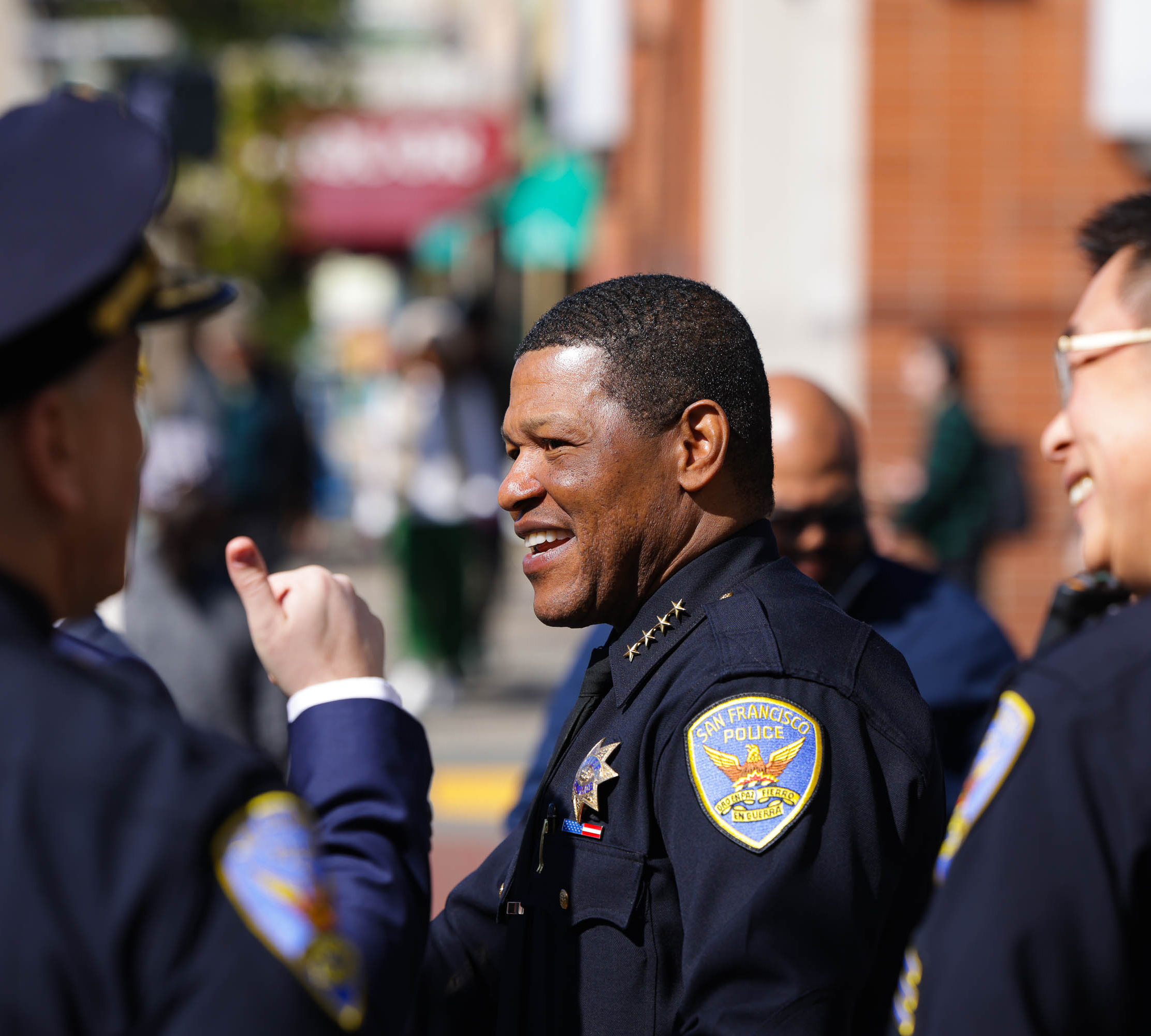 The image shows smiling uniformed officers from the San Francisco Police, engaged in a light-hearted conversation outdoors. One officer appears to be gesturing animatedly.