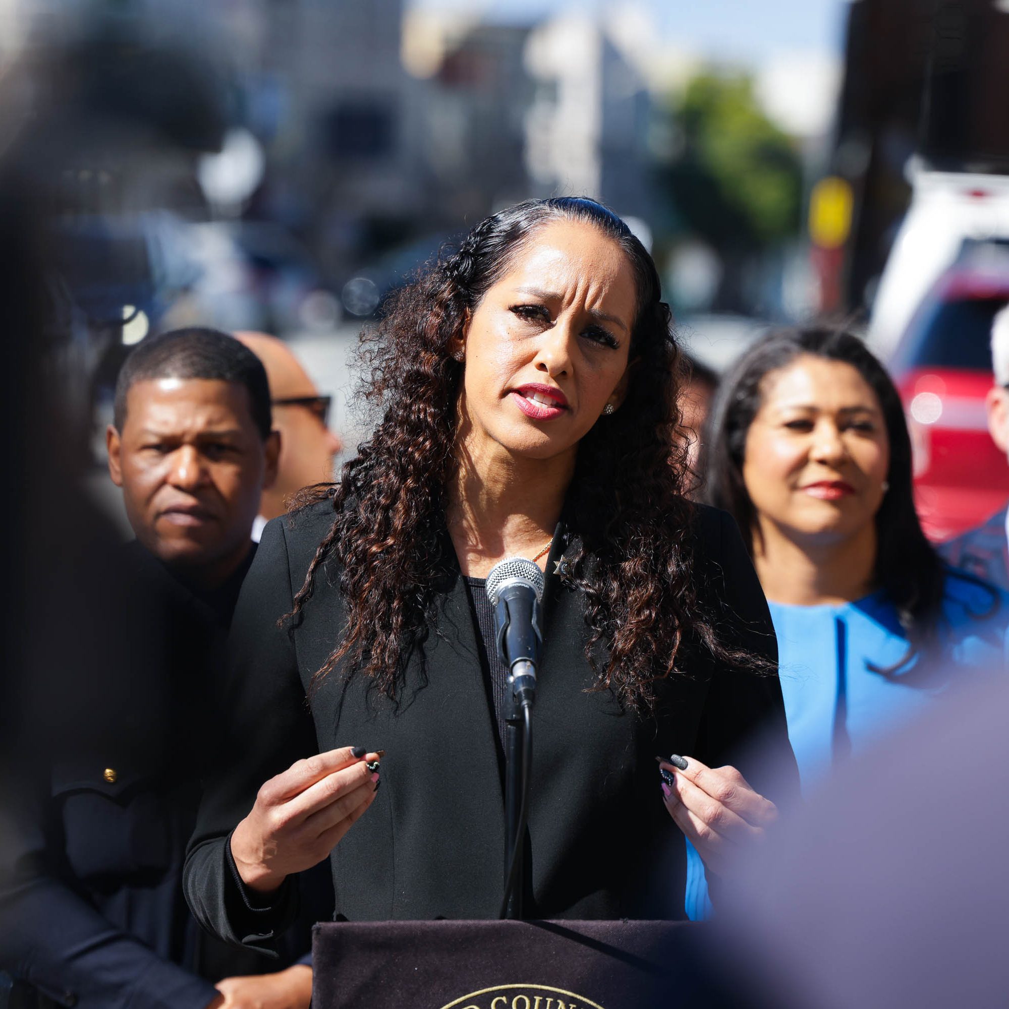 A woman with curly hair speaks passionately at a microphone outdoors, surrounded by people including a man in sunglasses and a woman in a blue outfit.