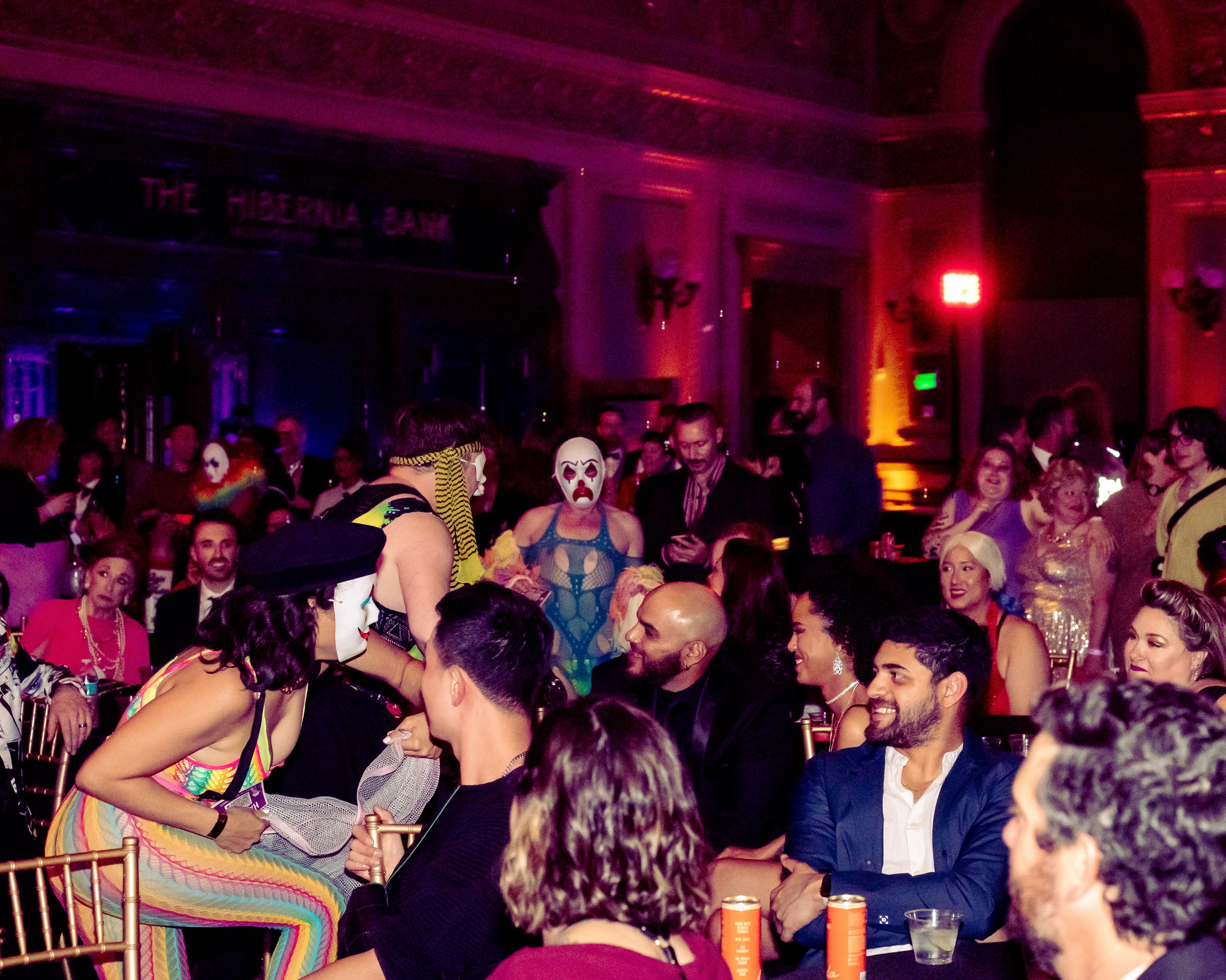 A lively crowd at a party with people in colorful outfits and face paint, seated and standing in a dimly lit room with ornate decor and an atmosphere of excitement.