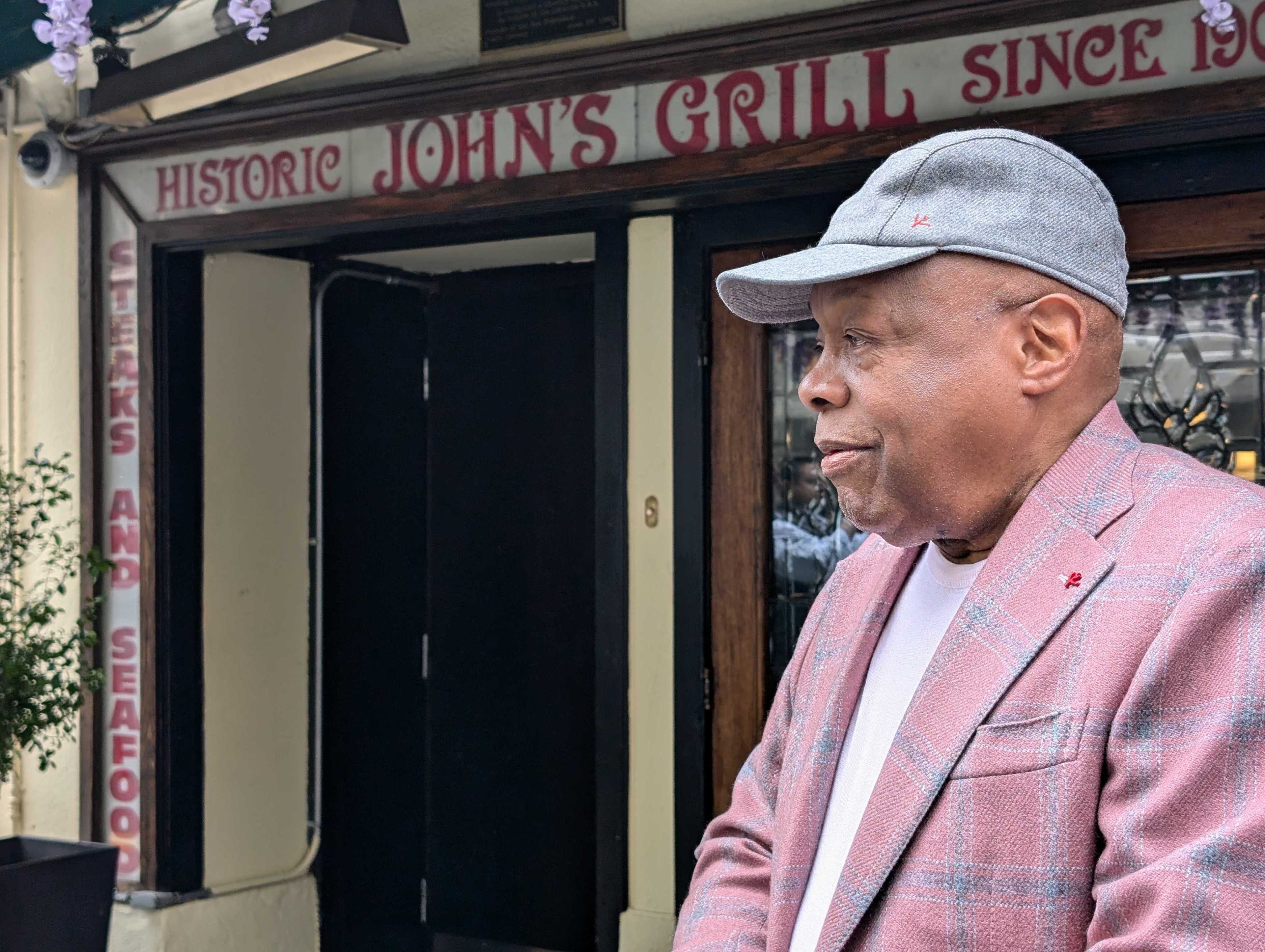 A man in a gray cap and pink plaid blazer stands in front of &quot;John's Grill,&quot; a historic steak and seafood restaurant, with the sign visible behind him.