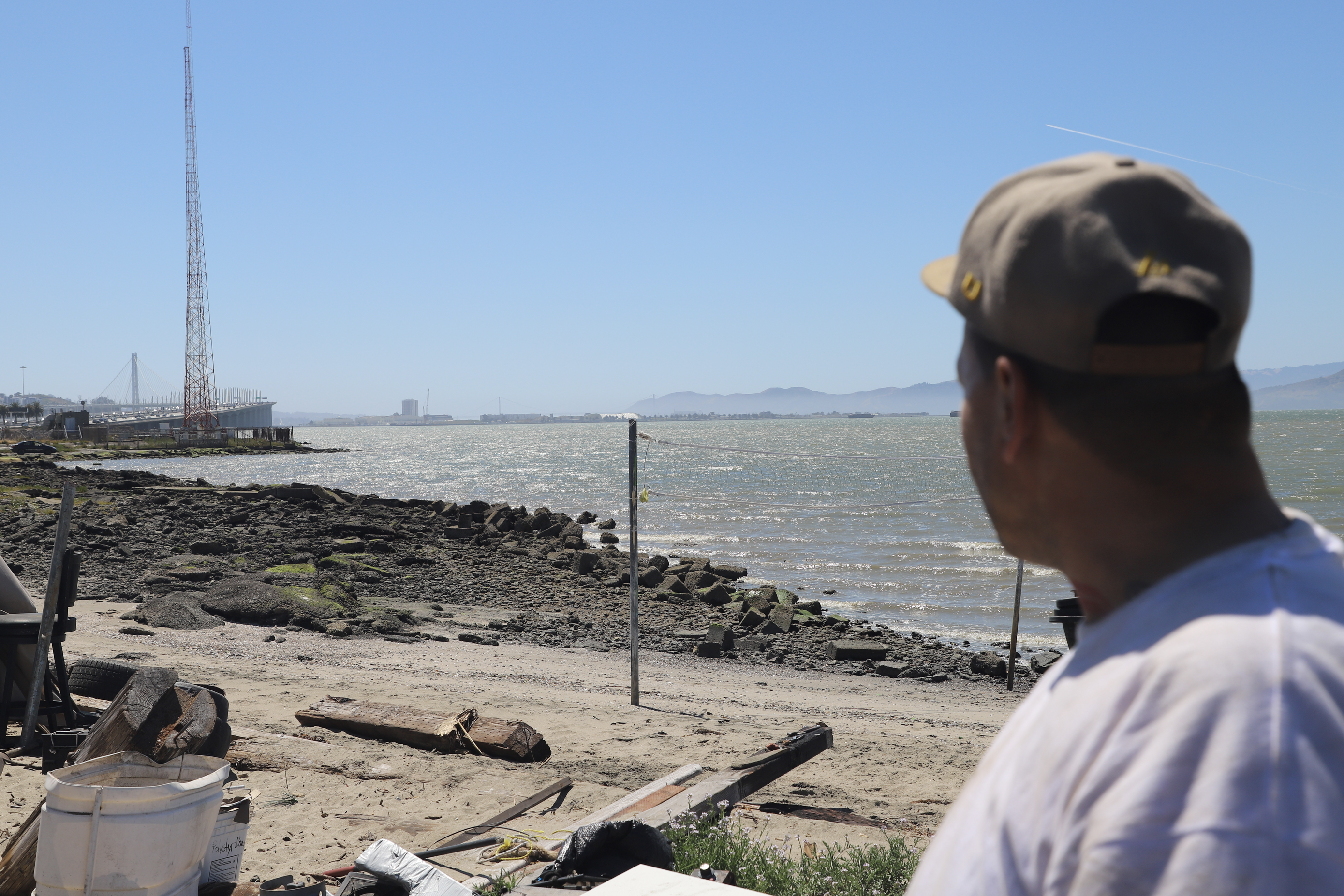 A person in a beige cap looks out over a rocky shoreline with calm waters, distant bridges, a tall red tower, and a clear blue sky in the background.