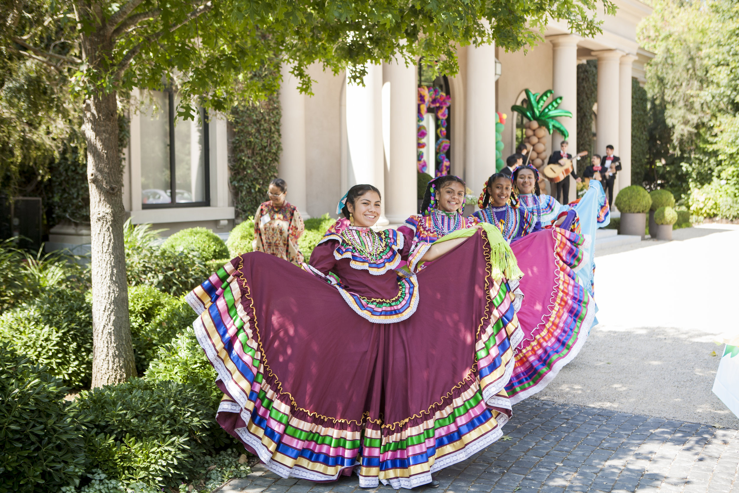 Women in colorful traditional dresses are dancing outdoors near a building adorned with greenery and columns, while musicians play in the background.