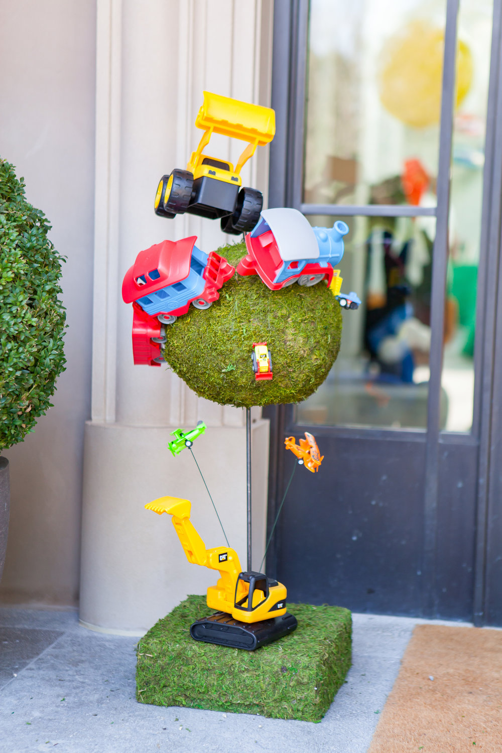 Toy vehicles, including a yellow bulldozer, red and blue trucks, and small planes, are creatively arranged on and around a moss-covered sphere.