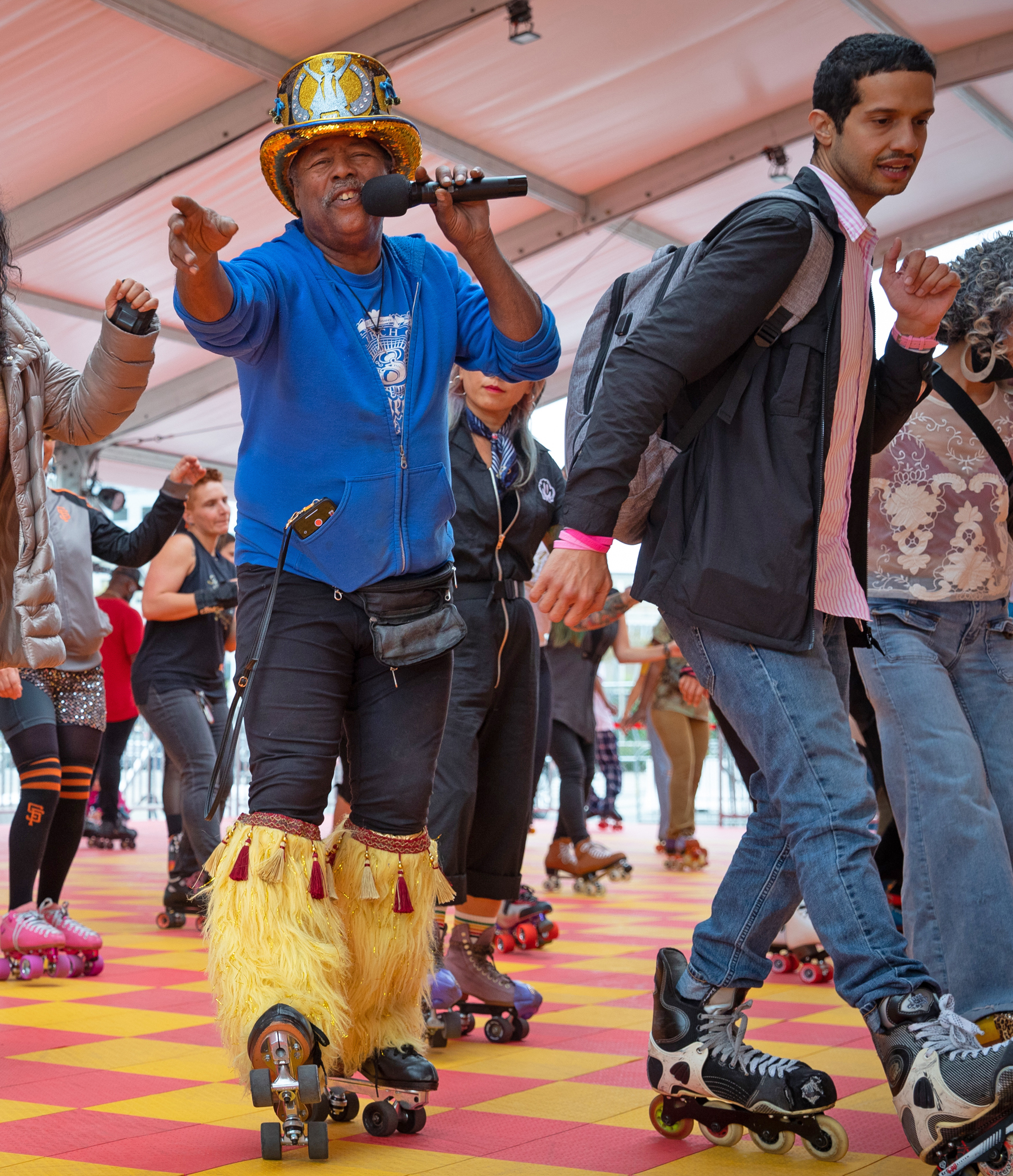 A man wearing a decorated hat, blue hoodie, and yellow furry leg warmers sings into a microphone while roller-skating. Other people are roller-skating around him.