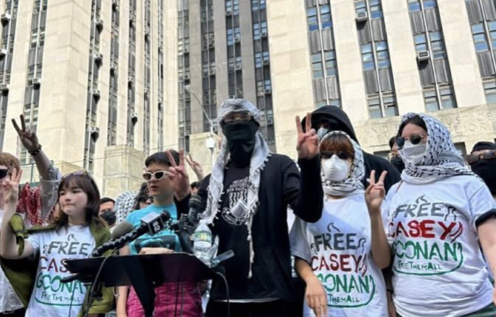 A group of people, some wearing masks and headscarves, stand in front of a tall building holding up peace signs and wearing &quot;Free Casey Conan&quot; shirts.