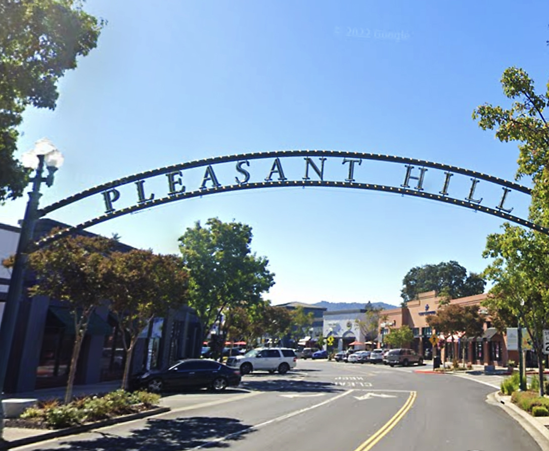 The image shows a street scene under a large metal arch reading &quot;PLEASANT HILL,&quot; with trees, parked cars, buildings, and a clear blue sky in the background.
