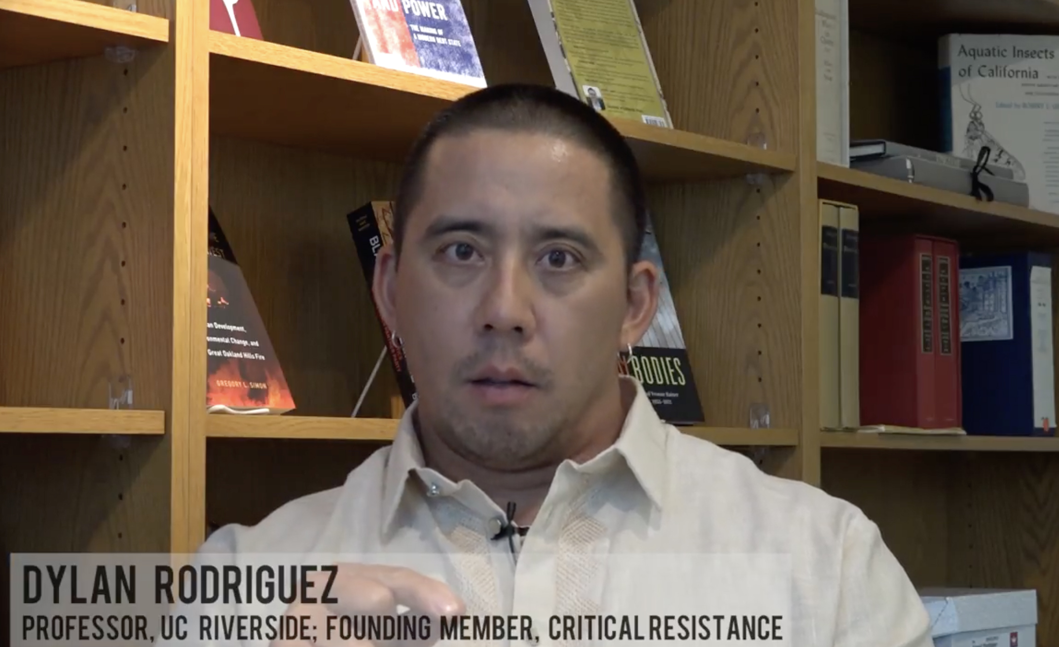 A man in a beige shirt stands in front of a wooden bookshelf filled with various books. The caption reads: &quot;Dylan Rodriguez, Professor, UC Riverside; Founding Member, Critical Resistance.&quot;