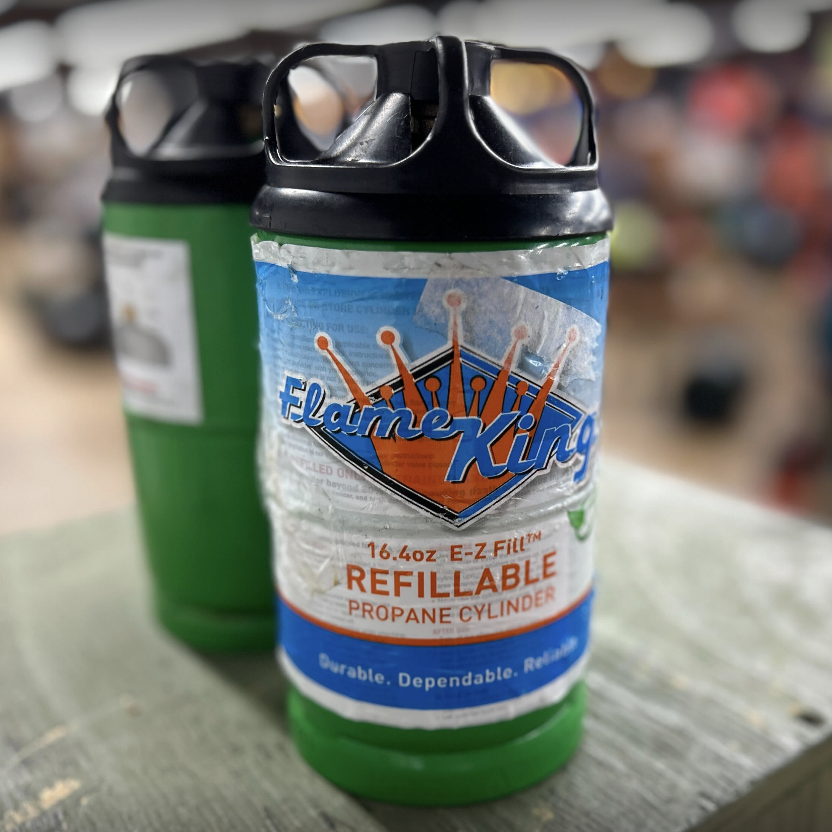 The image shows two green Flame King refillable propane cylinders with black caps, standing close together on a wooden surface in a blurred indoor setting.