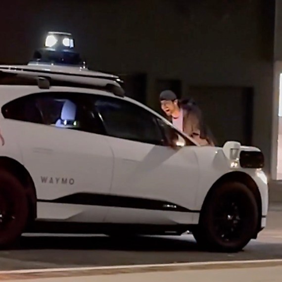 A white Waymo autonomous vehicle with a roof sensor is parked on the street at night, and a person is standing next to the driver's window, looking inside.