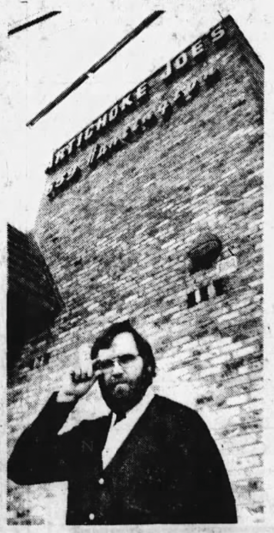 The image shows a person standing in front of a tall brick building labeled &quot;Artichoke Joe's.&quot; The person is saluting with their right hand, wearing glasses and a dark jacket.