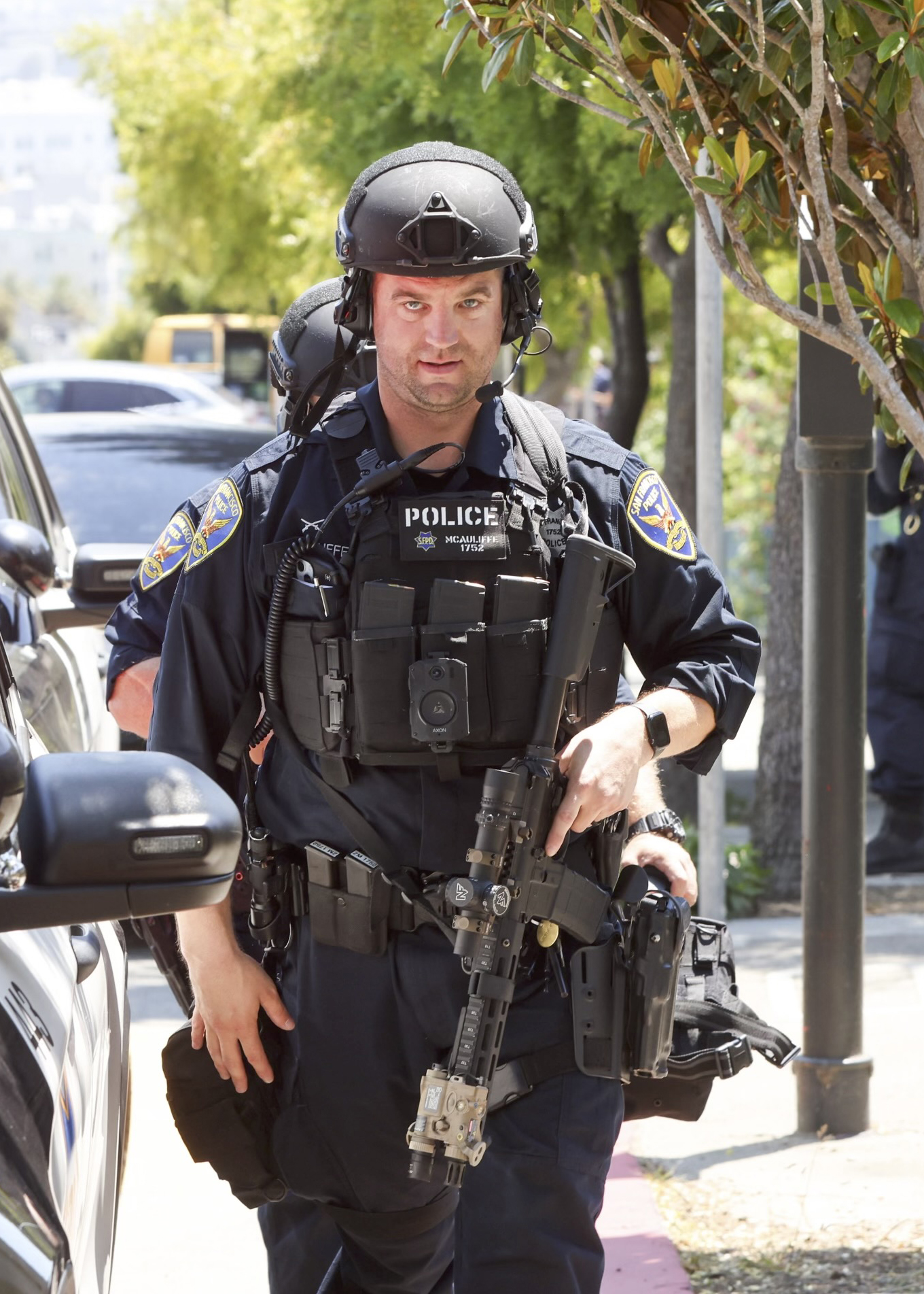 The image shows a police officer in tactical gear, including a helmet, body armor, and a rifle, standing near a police vehicle on a sunny day. Trees are visible in the background.